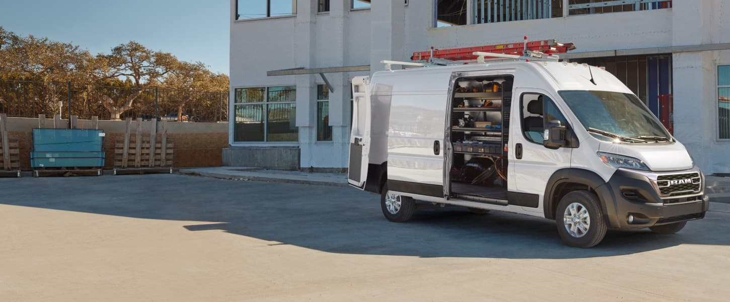 A passenger-side front angle of a white 2026 Ram ProMaster 3500 SLT Plus Cargo Van High Roof parked beside a commercial building with a ladder on its roof rack, its rear doors and sliding side door open revealing shelving full of tools in the cargo area.