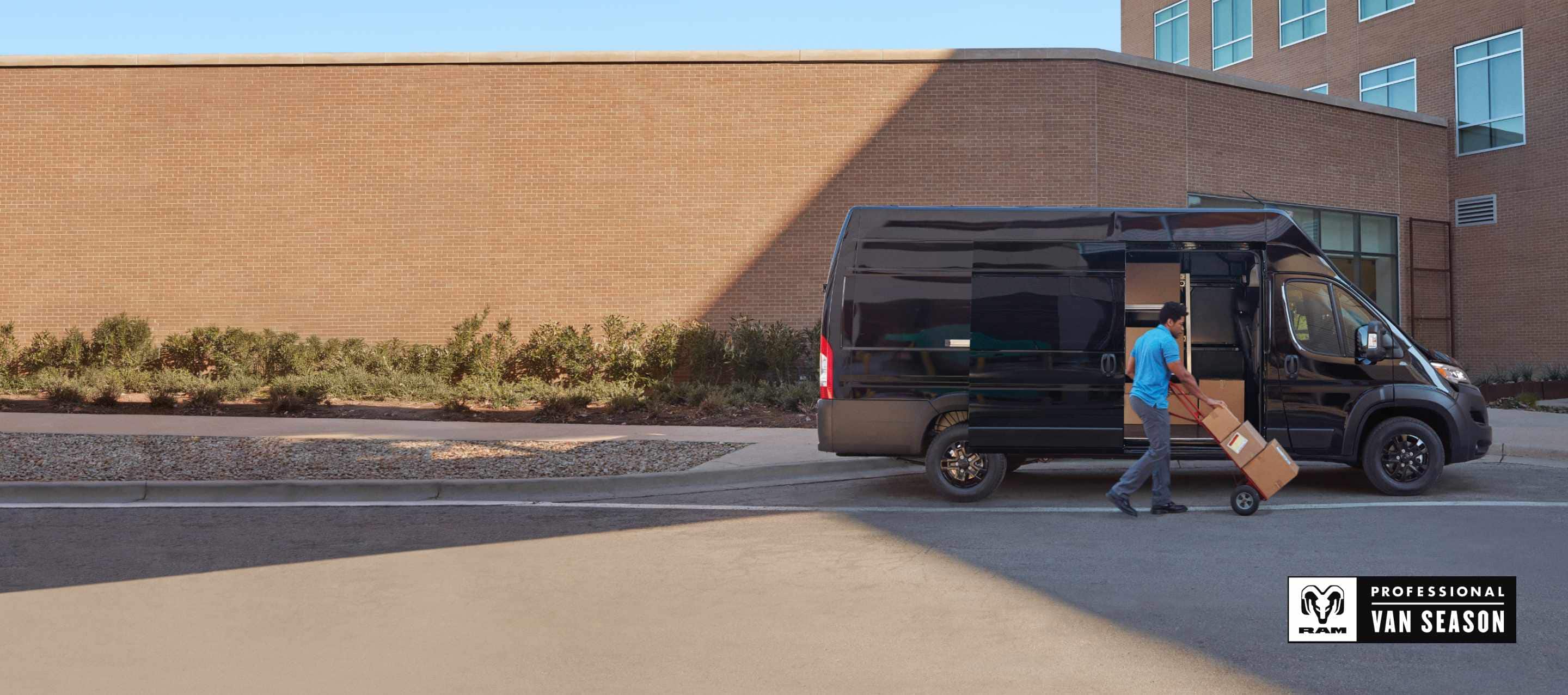 A man pushing packages on a dolly near the open passenger-side sliding door on a black 2025 Ram ProMaster 3500 SLT Plus Super High Roof Cargo Van with the Black Appearance Package. Ram Professional Van Season.