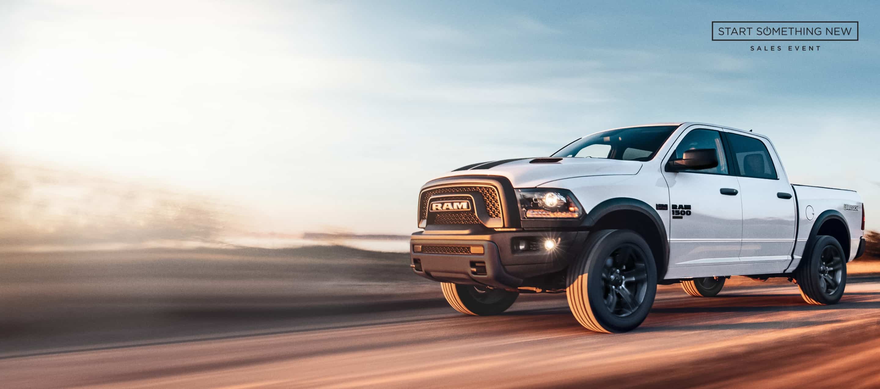 A white 2024 Ram 1500 Classic Warlock 4x4 Crew Cab traveling down a highway. The Start  Something New Sales Event.