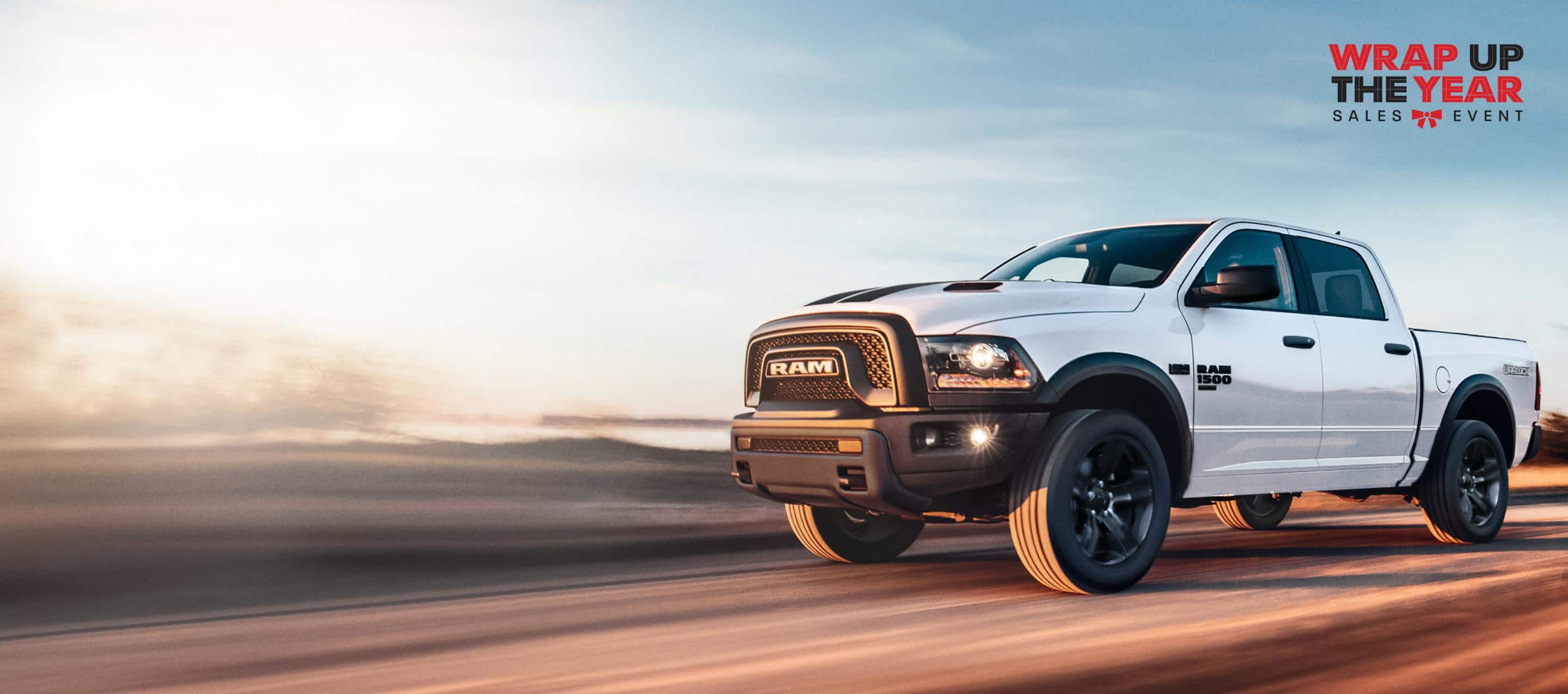 A white 2024 Ram 1500 Classic Warlock 4x4 Crew Cab traveling down a highway. The Wrap Up The Year Sales Event.