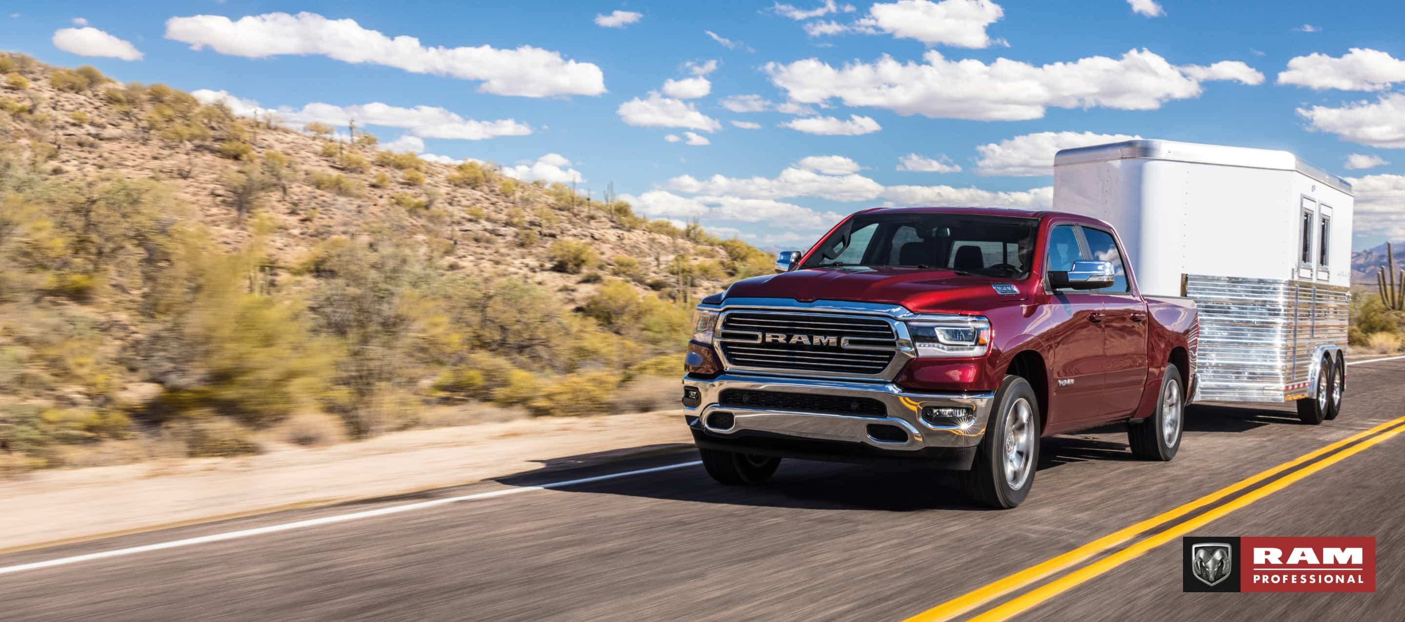 A red 2024 Ram 1500 Laramie Crew Cab towing a horse trailer as it is driven down a highway in the desert. Ram Professional.