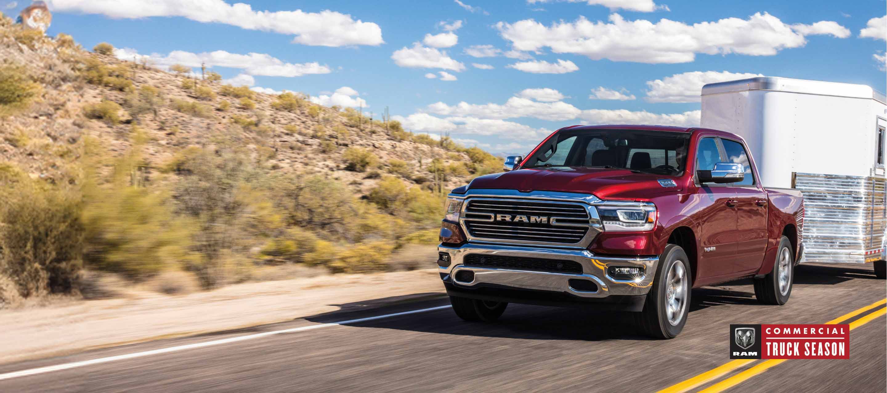 A red 2024 Ram 1500 Laramie Crew Cab towing a horse trailer as it is driven down a highway in the desert. Ram Commercial Truck Season.