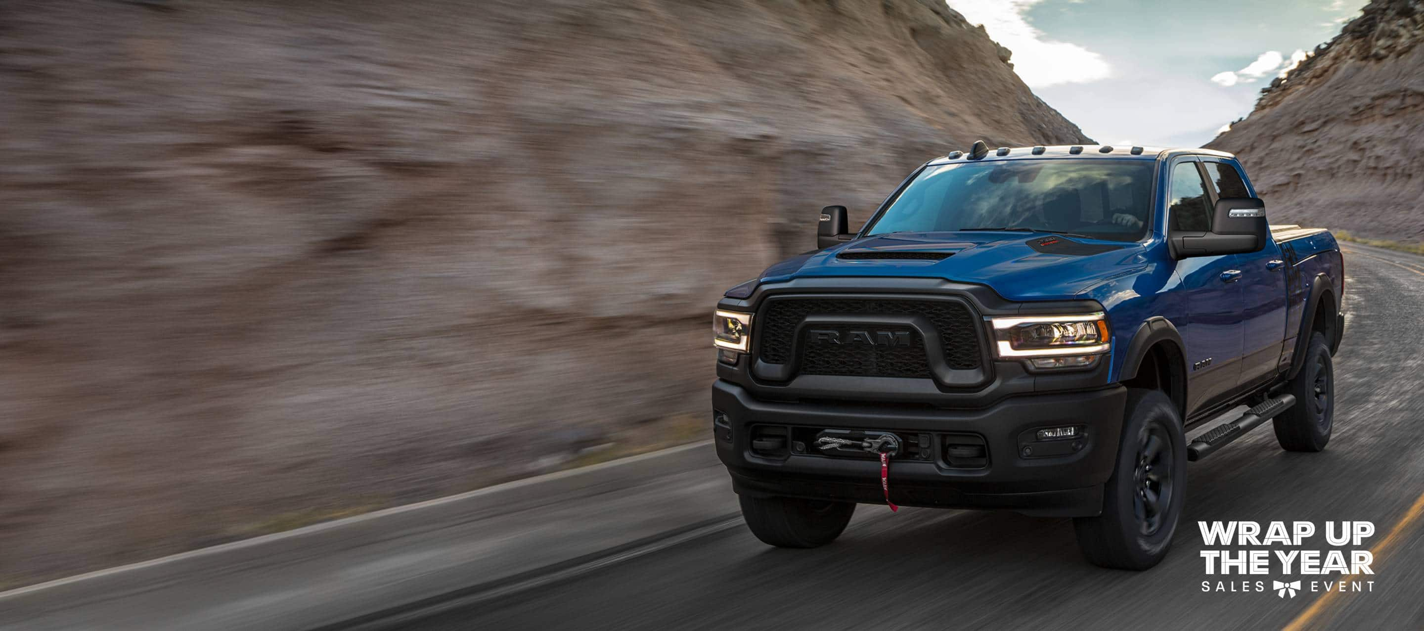 A blue 2024 Ram 2500 Power Wagon Crew Cab taking a curve on a mountain highway. The Wrap Up The Year Sales Event.