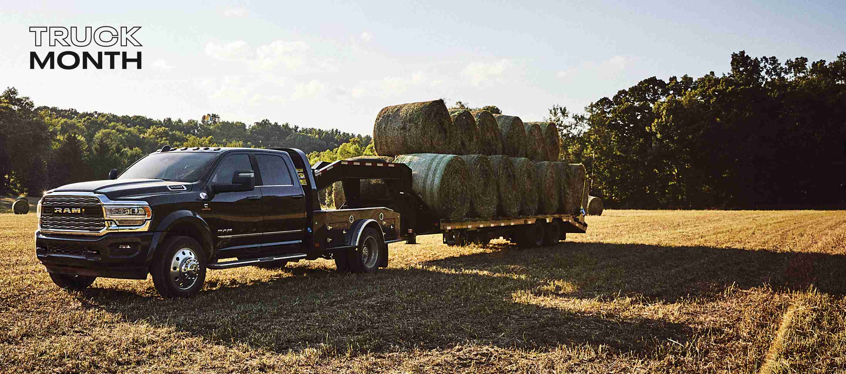 A black 2024 Ram 5500 Limited 4x4 Chassis Crew Cab with a gooseneck trailer loaded with several bales of hay, parked in an open field. Truck Month.