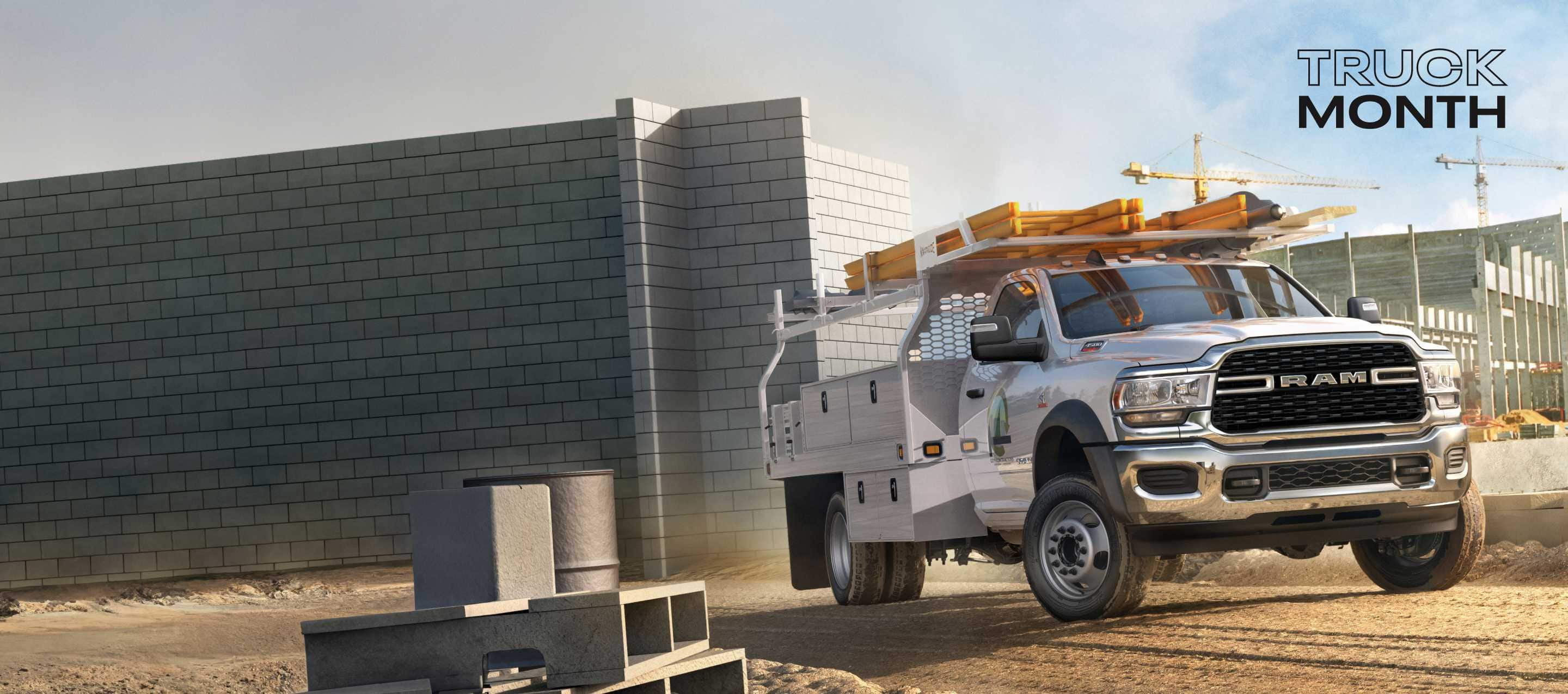 A white 2024 Ram 4500 Chassis Cab Regular Cab with a utility upfit, parked at a large commercial construction site. Truck Month Sales Event.