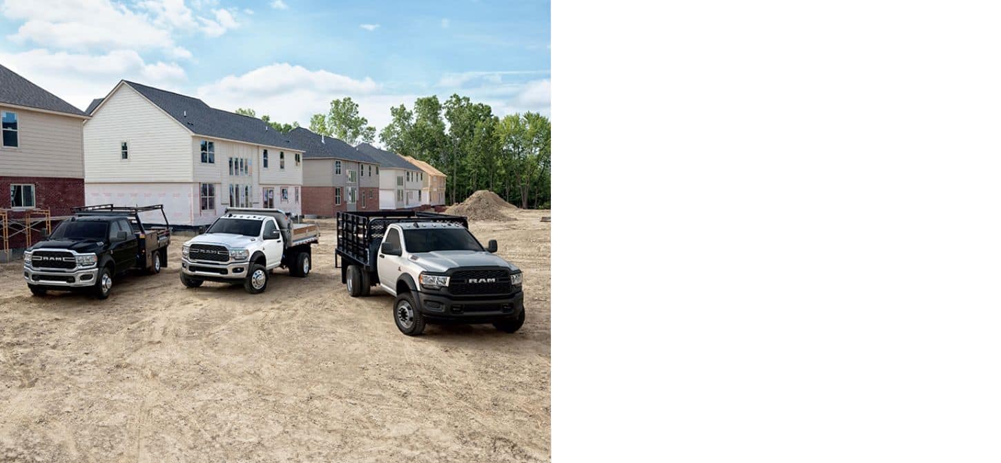 Three 2023 Ram Chassis Cabs parked at a construction site, each with a different upfit. From left to right: a black Ram 5500 Tradesman Chassis Cab with a stake bed upfit, a white Ram 5500 SLT Chassis Cab with dump upfit and a white Ram 4500 Tradesman Chassis Cab with landscaper upfit.