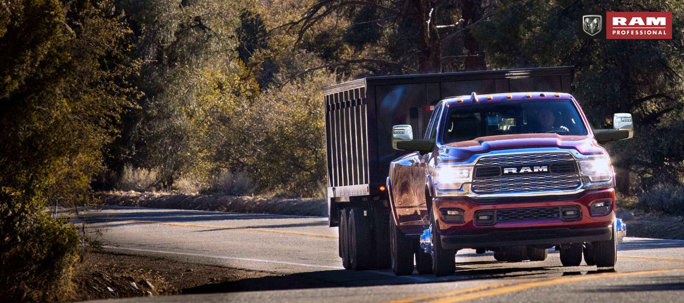 A red 2024 Ram 3500 4x4 Crew Cab traveling on a highway in the woods towing a dump body trailer. Ram Professional.