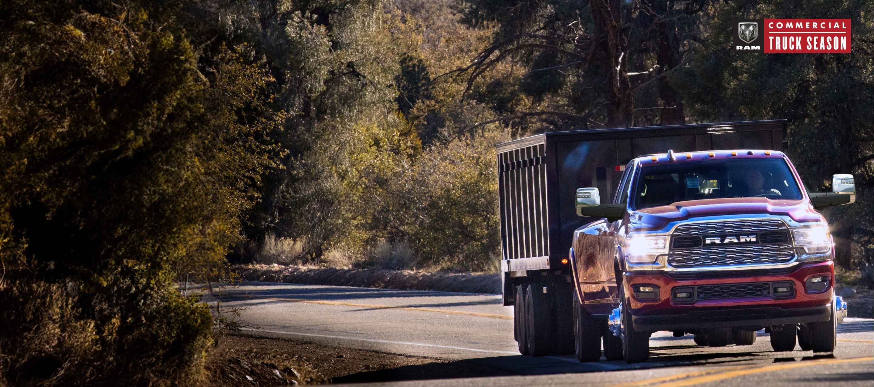 A red 2024 Ram 3500 4x4 Crew Cab traveling on a highway in the woods towing a dump body trailer. Ram Professional.