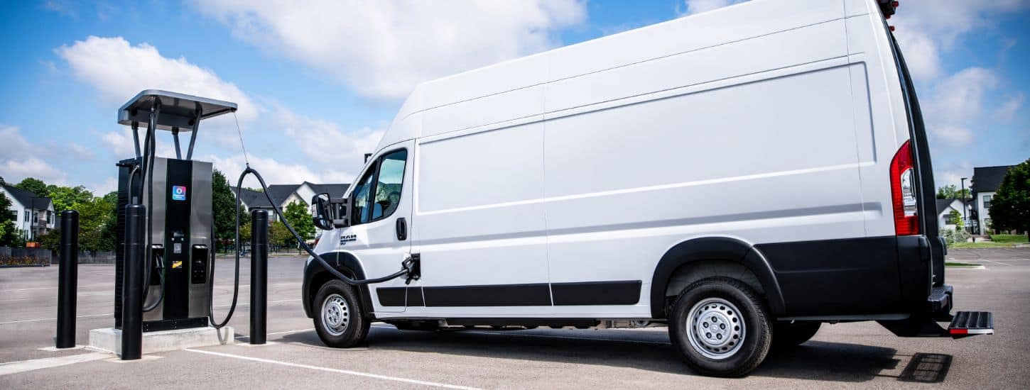 A white 2024 Ram ProMaster EV 3500 Step Van parked beside a Free2move Charge charging station with its charging cord plugged into the Ram ProMaster.