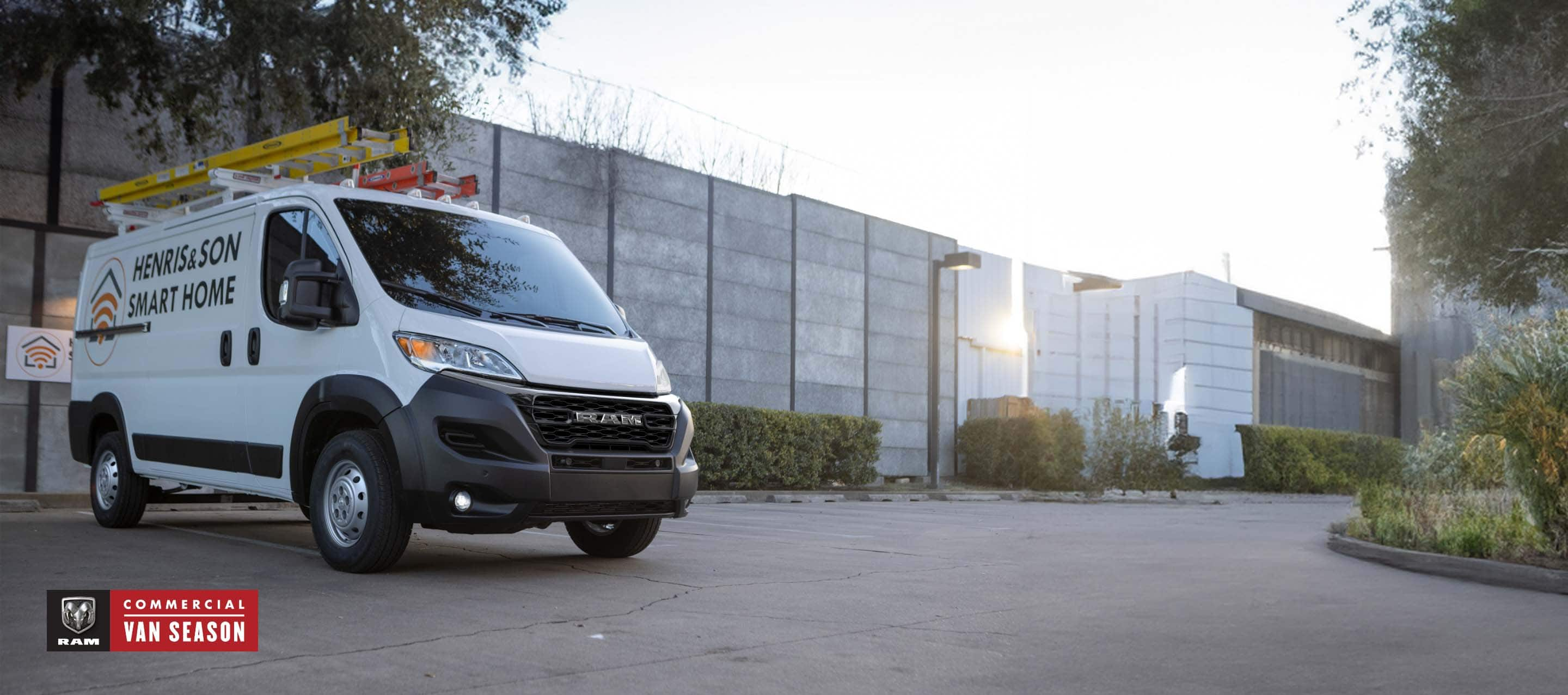 A white 2024 Ram ProMaster 1500 Tradesman Standard Roof Cargo Van with two ladders on its roof rack and the logo of a smart home installation company on its side panel, parked beside a commercial building. Ram Commercial Van Season.