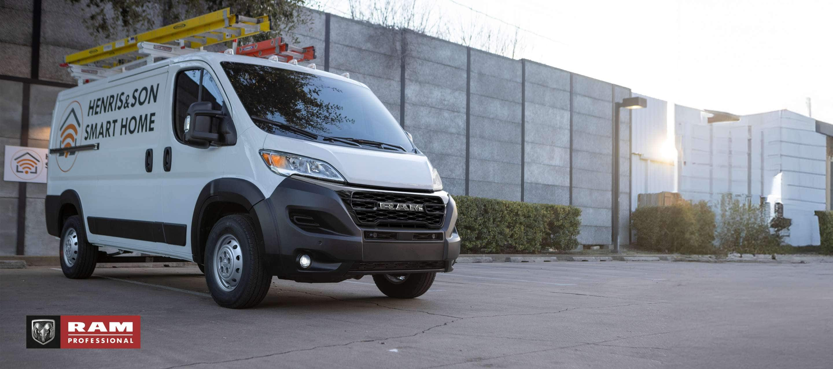 A white 2024 Ram ProMaster 1500 Tradesman Standard Roof Cargo Van with two ladders on its roof rack and the logo of a smart home installation company on its side panel, parked beside a commercial building. Ram Professional.