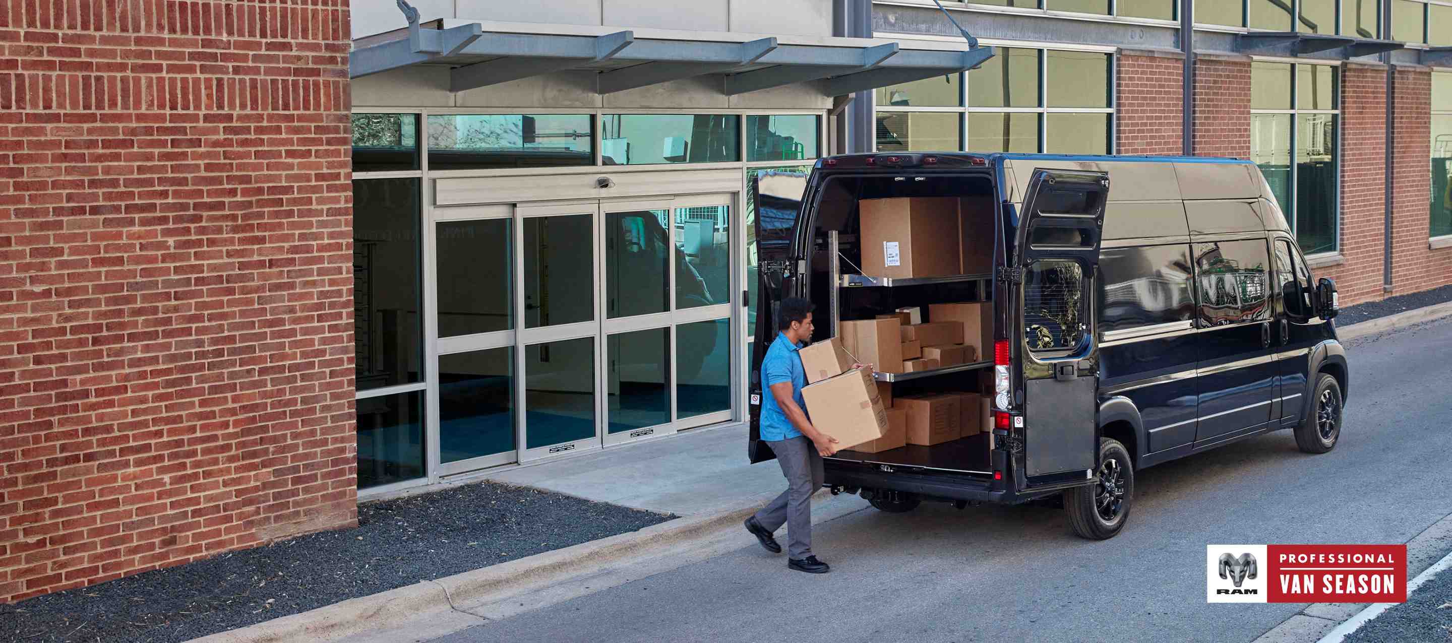 A man with boxes approaching the open rear doors of a black 2024 Ram ProMaster 3500 SLT Plus Cargo Van Super High Roof with the Black Appearance Package. Professional Van Season.