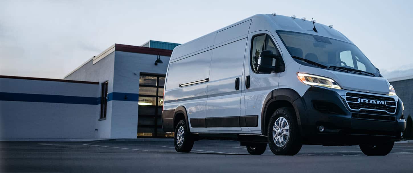 A passenger-side front angle of a white Ram ProMaster EV 3500 SLT High Roof Cargo Van with its headlamps on, parked in the lot of a commercial building.