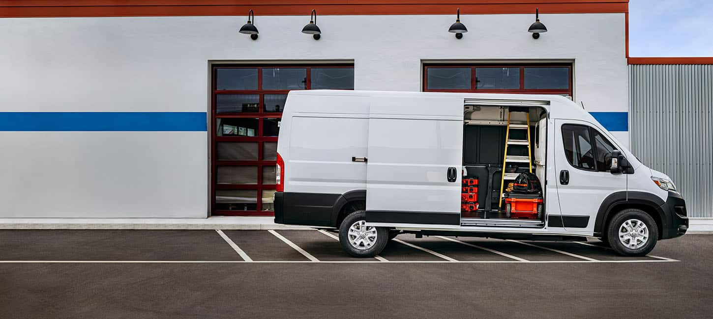 A passenger-side profile of a white Ram ProMaster EV 3500 SLT High Roof Cargo Van parked beside a commercial garage with its sliding side door open, revealing tools, equipment and a ladder inside.