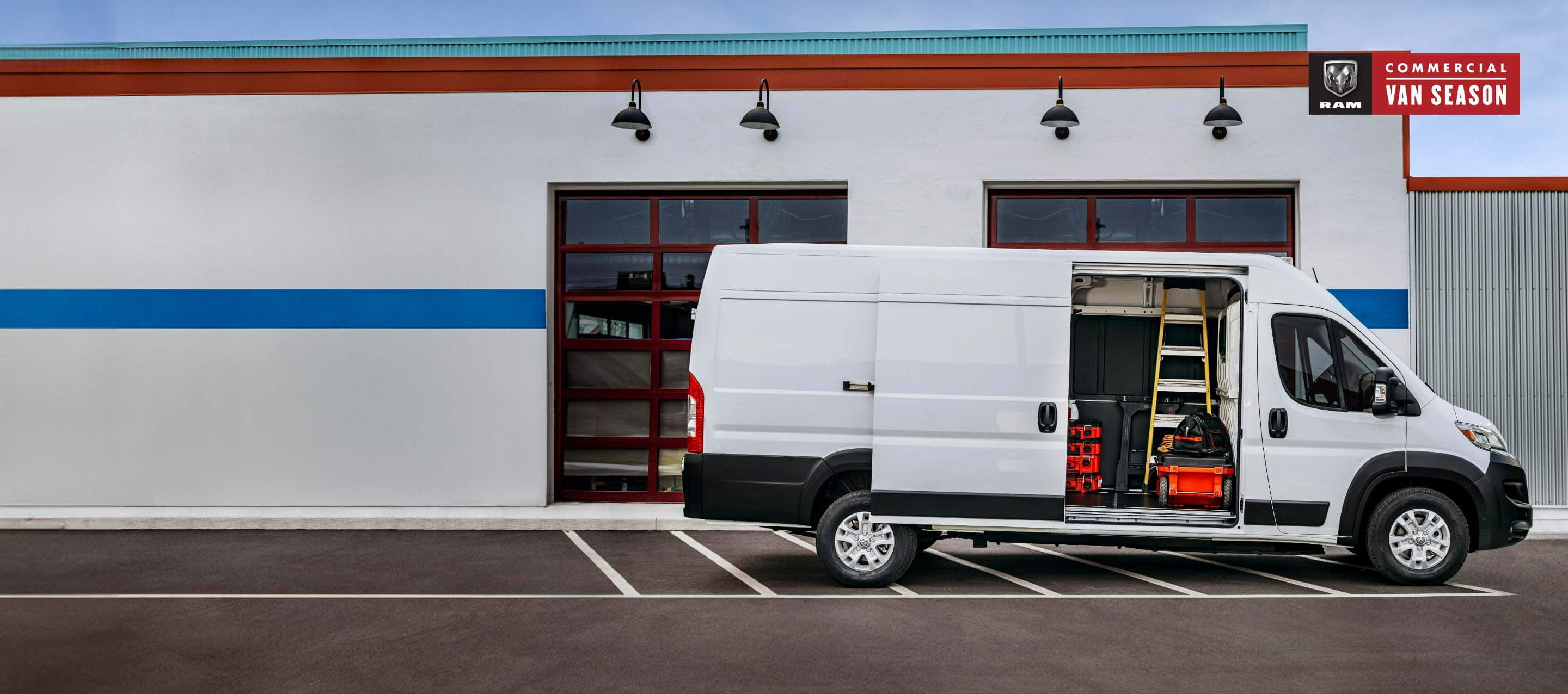 A passenger-side profile of a white 2024 Ram ProMaster EV High Roof Cargo Van (Preproduction Cargo Van shown. Actual production vehicle and feature may vary), parked beside a commercial garage with its passenger-side sliding door open revealing a ladder, tools and equipment inside. Ram Commercial Van Season.