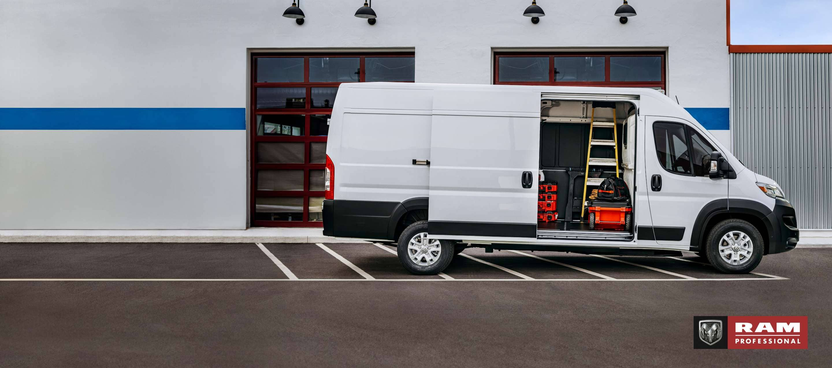 A passenger-side profile of a white 2024 Ram ProMaster EV High Roof Cargo Van (Preproduction Cargo Van shown. Actual production vehicle and feature may vary), parked beside a commercial garage with its passenger-side sliding door open revealing a ladder, tools and equipment inside. Ram Professional.