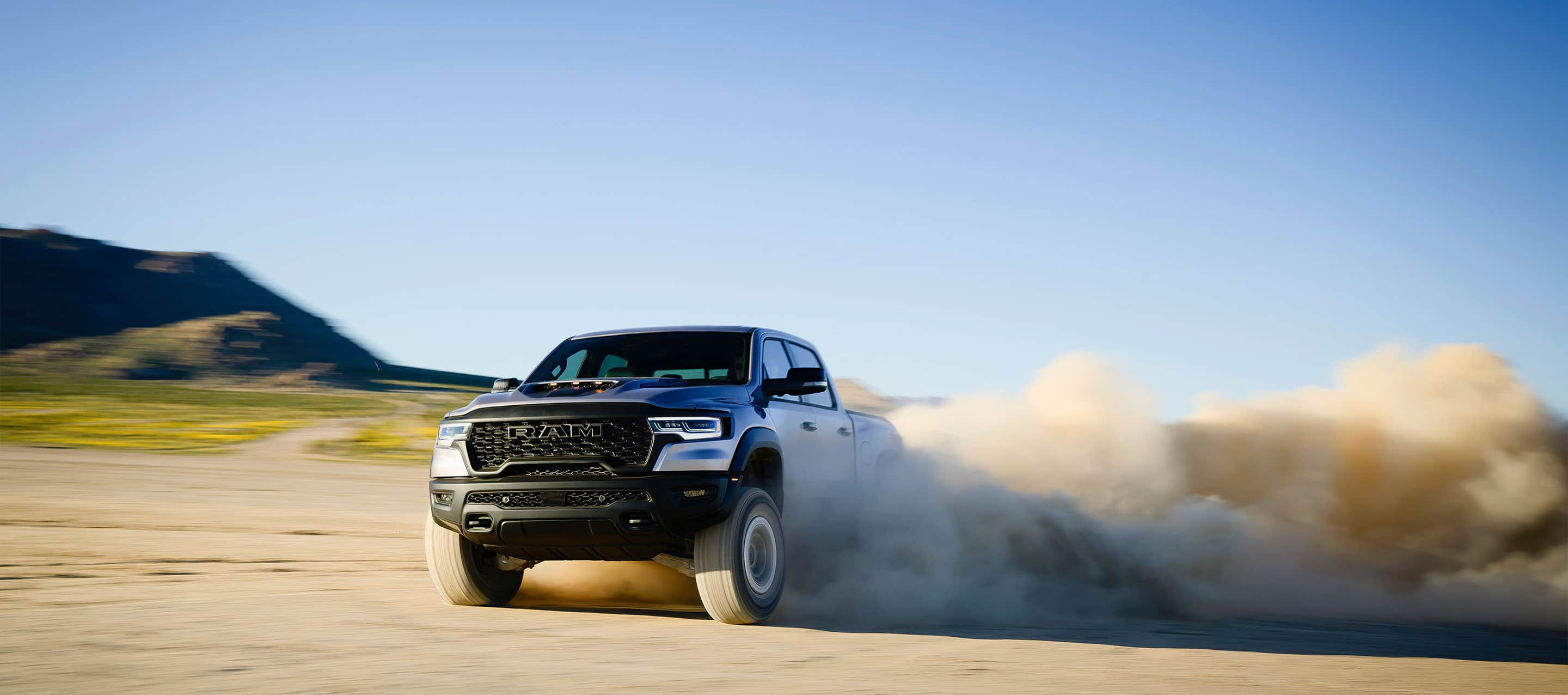 A 2025 Ram 1500 RHO Crew Cab being driven on sand as it kicks up dust in its wake. The background is blurred to indicate the truck is in motion.