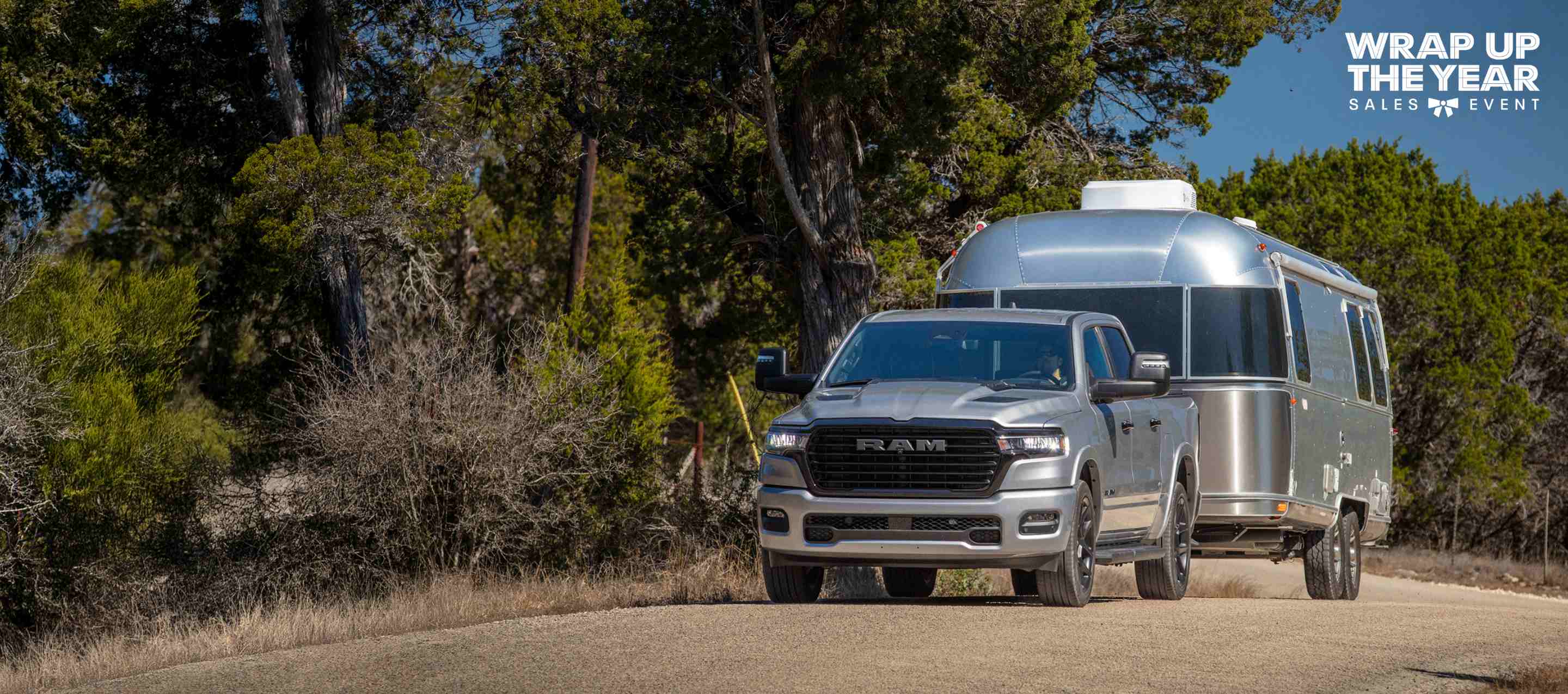 A silver 2025 Ram 1500 Laramie Crew Cab towing a large travel trailer as it's driven down a highway. The Wrap Up The Year Sales Event.