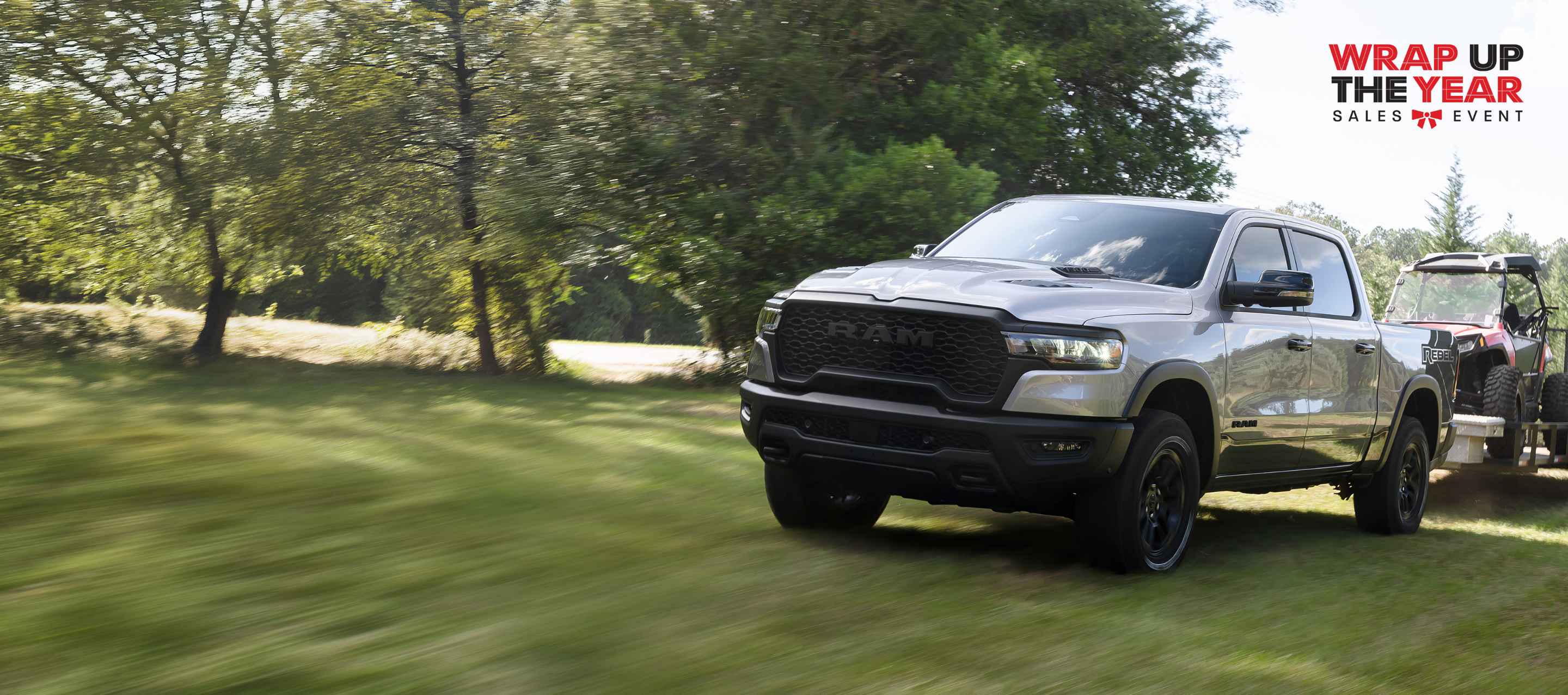 A silver 2025 Ram 1500 Rebel 4x4 Crew Cab traveling off-road, towing an ATV. The Wrap Up The Year Sales Event.