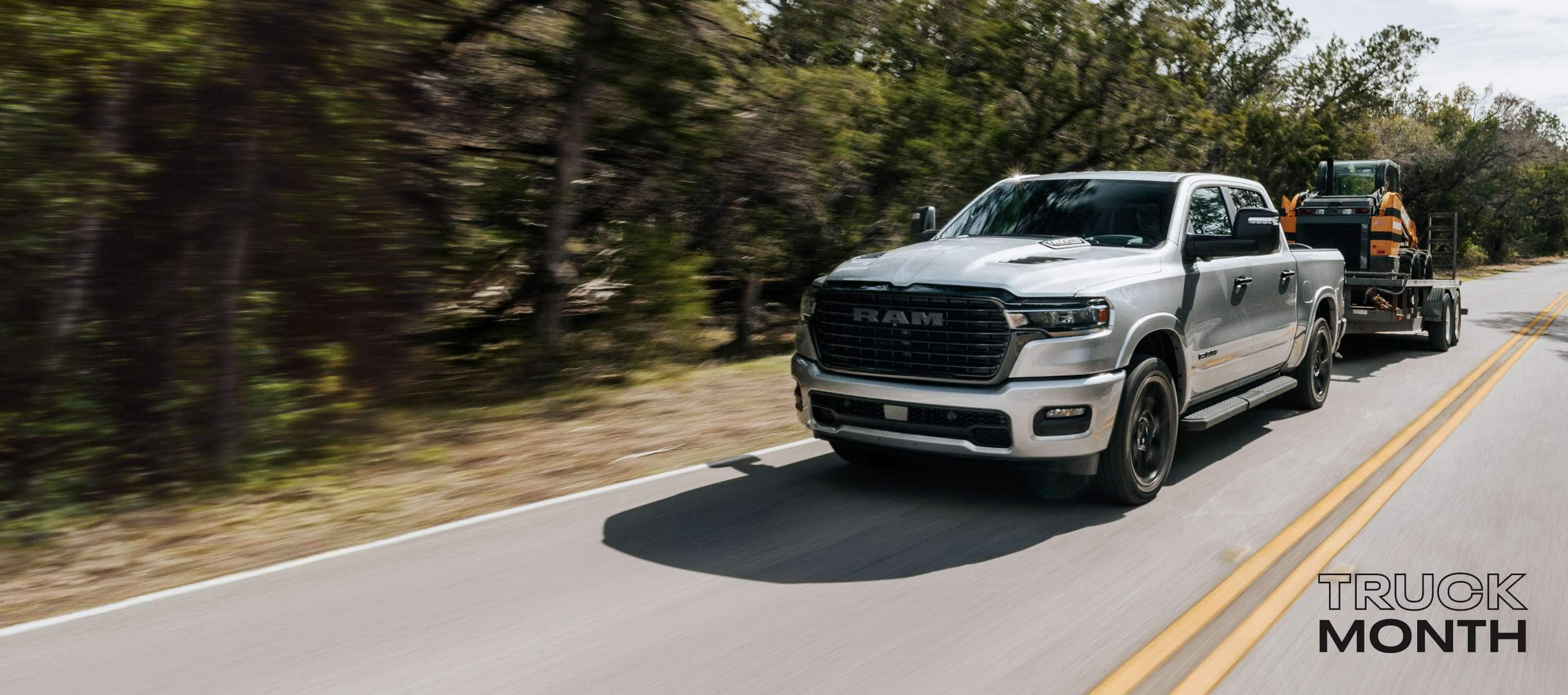 A silver 2025 Ram 1500 Laramie 4x4 Crew Cab traveling on a rural highway, towing earth-moving equipment on a tilt trailer. Truck Month Sales Event.