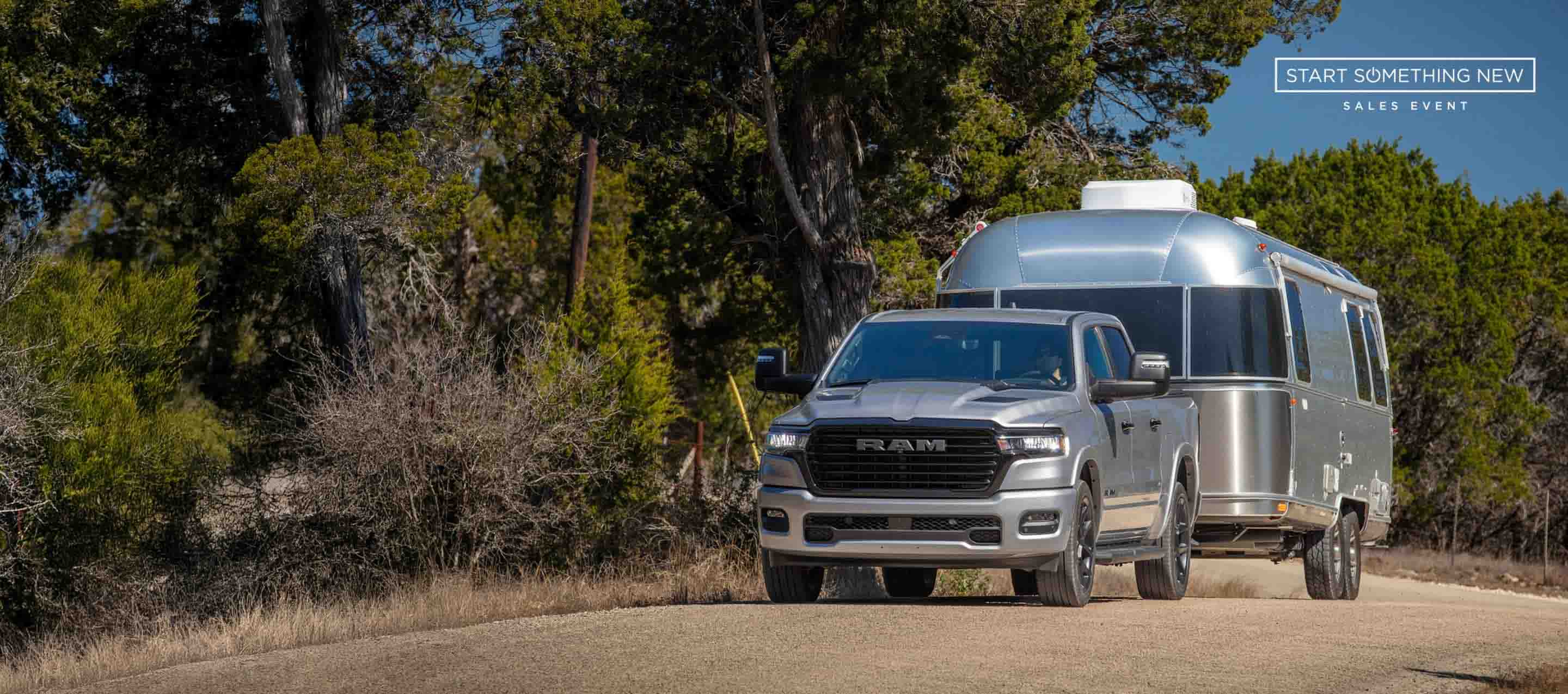 A silver 2025 Ram 1500 Laramie Crew Cab towing a large travel trailer as it's driven down a highway.  The Start  Something New Sales Event.