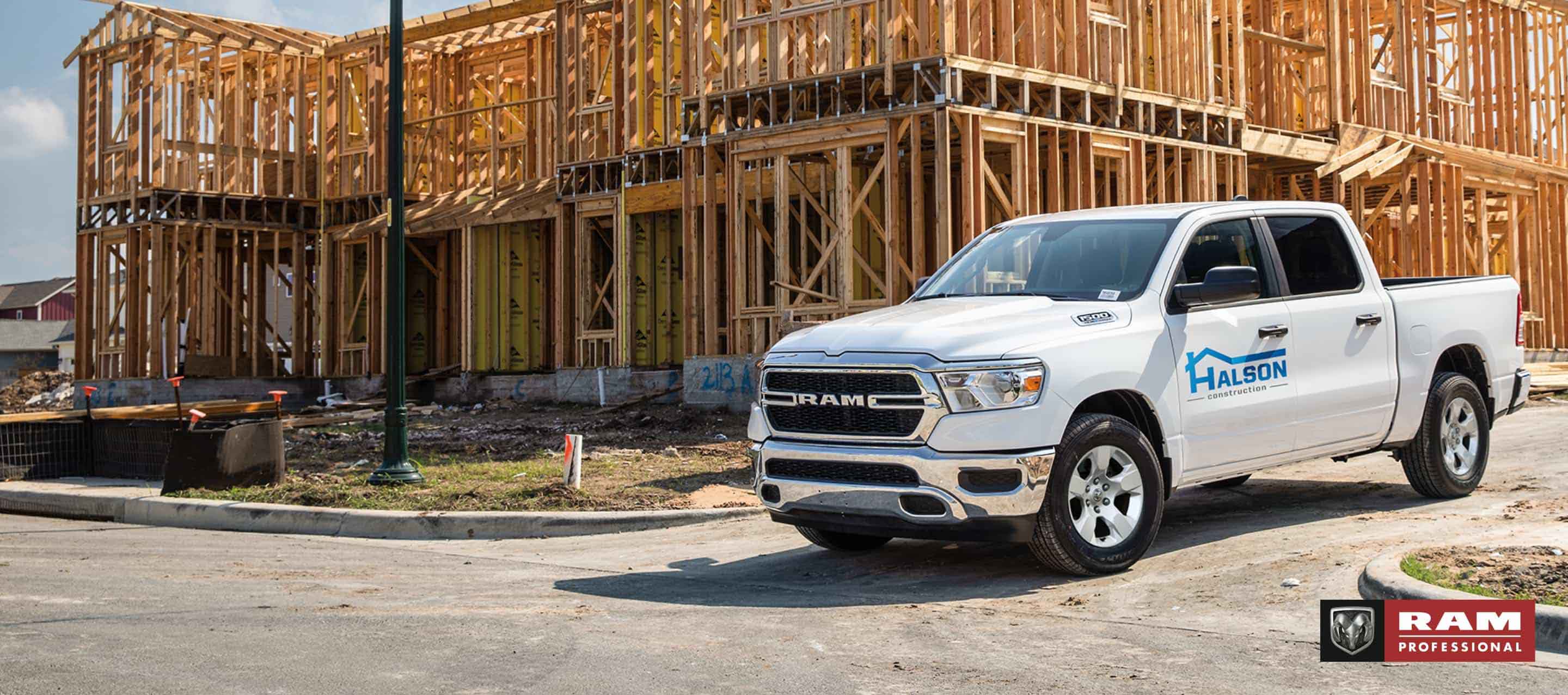 A white 2025 Ram 1500 Tradesman 4x4 Crew Cab, with construction company signage on its driver's door as it pulls away from a commercial building site. Ram Professional.