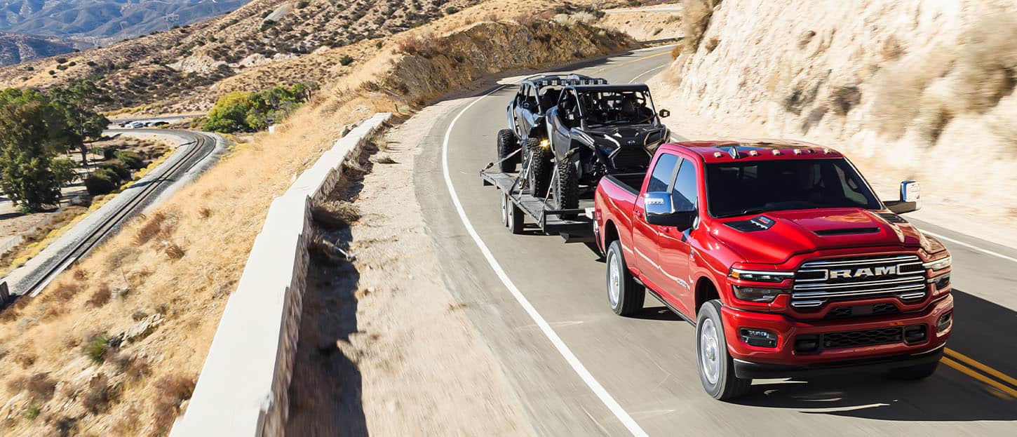 A red 2025 Ram 2500 Laramie 4x4 Crew Cab traveling on a highway in the mountains, while towing two ATVs.