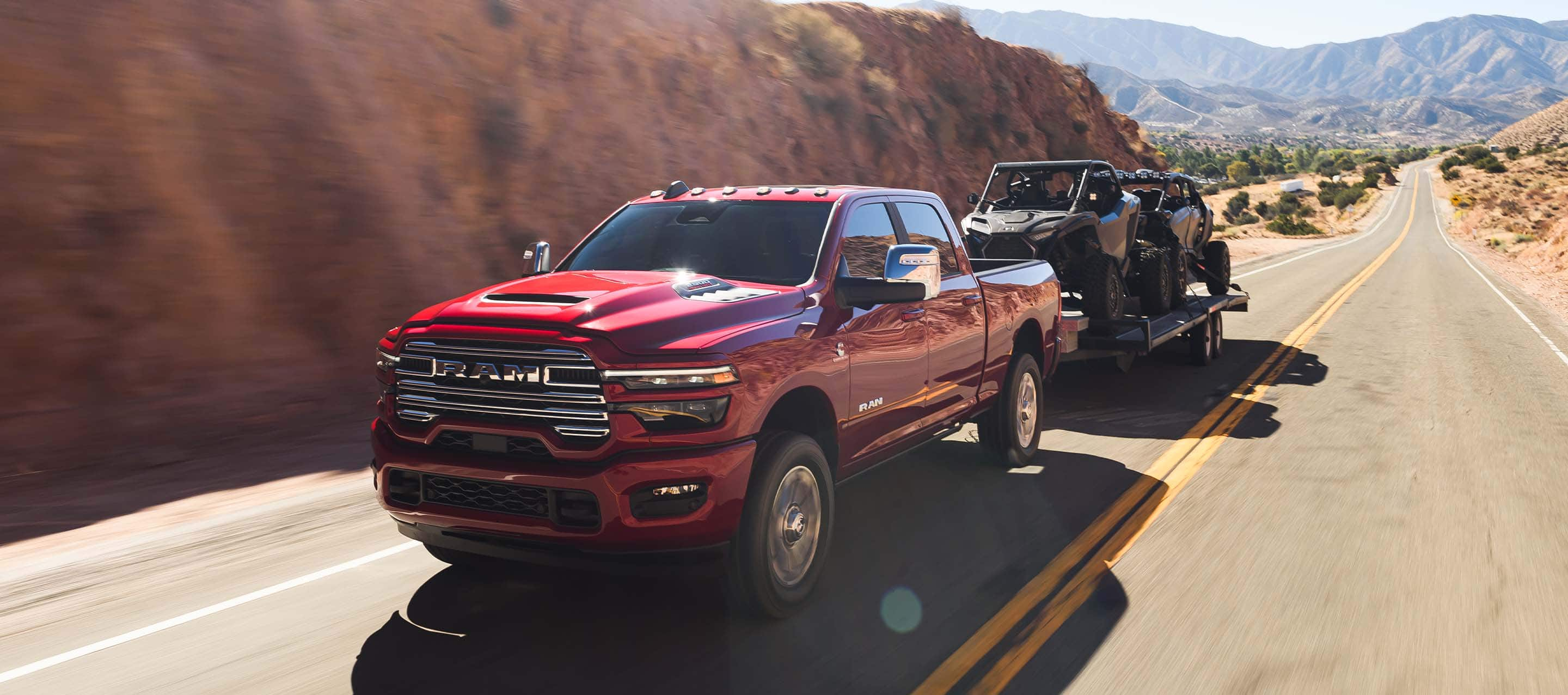 A red 2025 Ram 2500 Laramie 4x4 Crew Cab traveling on a highway in the mountains, while towing two ATVs.