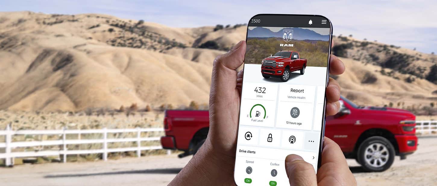 In the foreground a smartphone displaying a page from the Ram app, in the background a red 2025 Ram 2500 parked on a ranch.