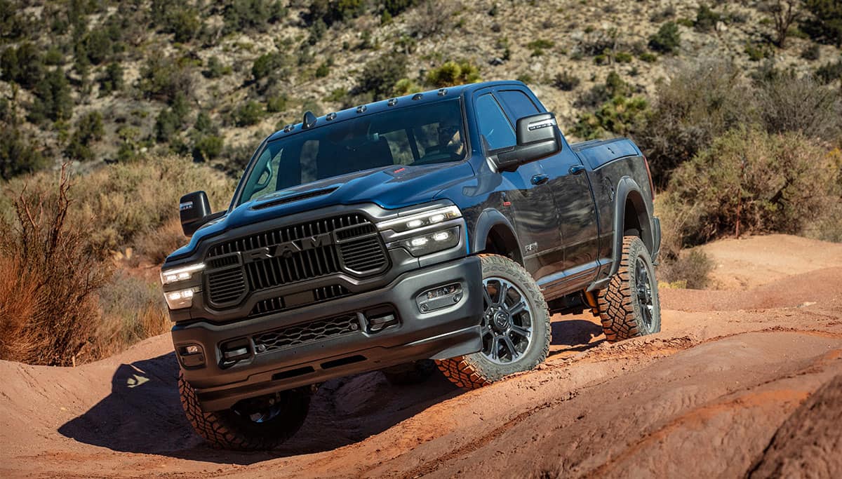 Display A black 2025 Ram 2500 Rebel 4x4 Crew Cab traveling off-road on uneven, rocky terrain.