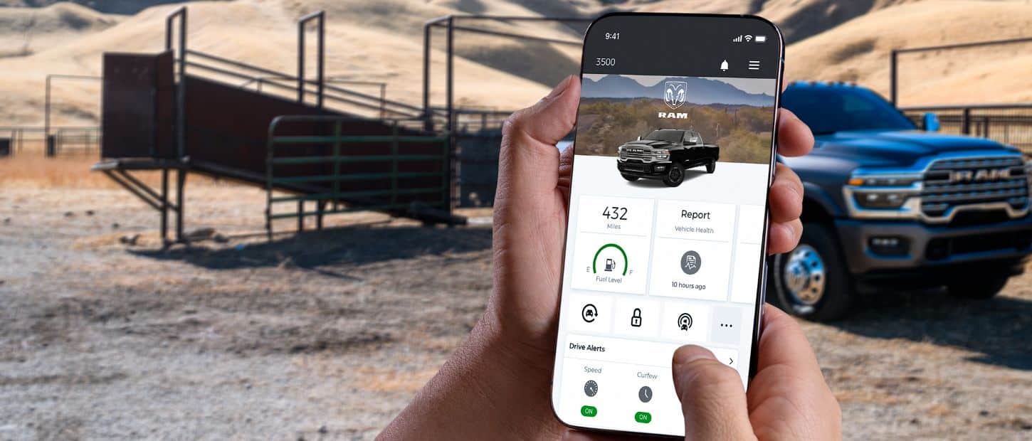 In the foreground a smartphone displaying a page from the Ram app, in the background a black 2025 Ram 3500 parked on a cattle ranch.