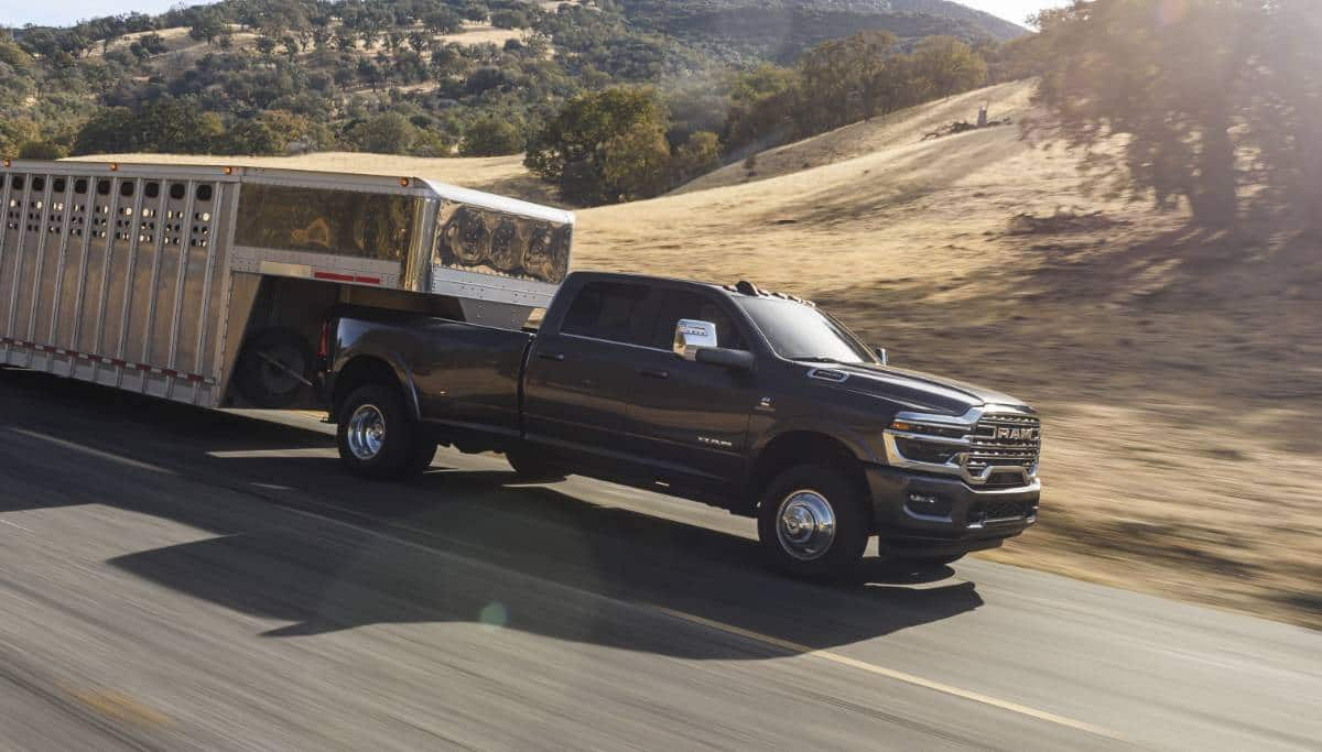 Display A 2025 Ram 3500 Limited Longhorn 4x4 Crew Cab towing a large fifth-wheel livestock trailer as it travels down a highway.