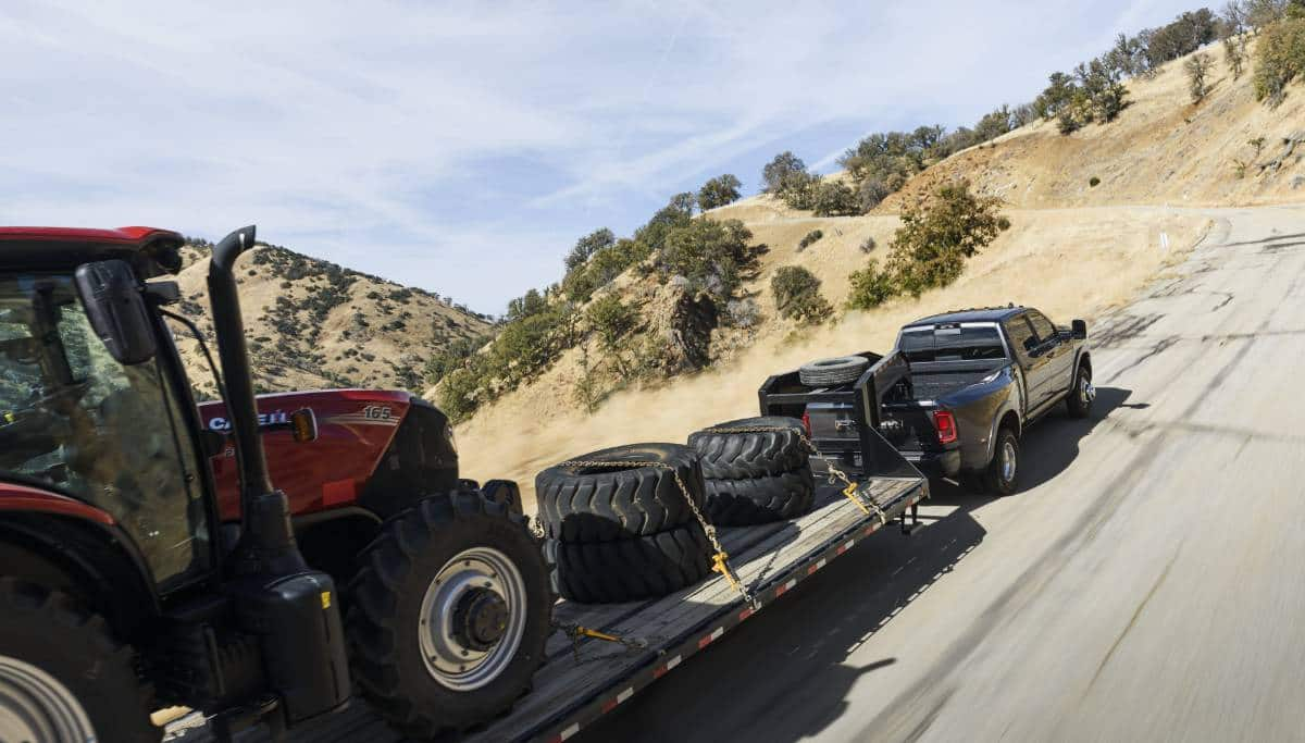 Display A 2025 Ram 3500 Limited Longhorn 4x4 Crew Cab towing a construction vehicle and four spare tires on a long fifth-wheel flatbed trailer as it travels on a mountain road.