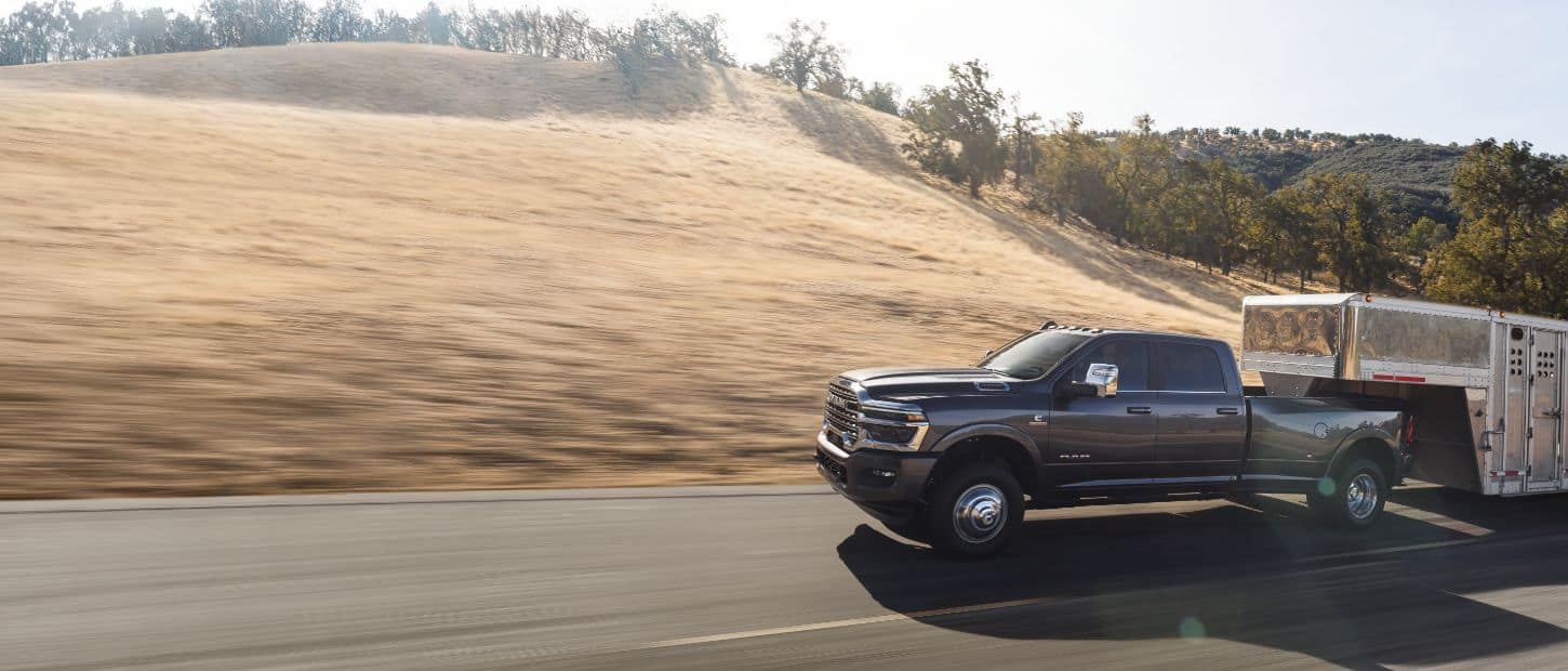 A gray 2025 Ram 3500 Limited Longhorn 4x4 Crew Cab towing a large fifth-wheel livestock trailer as it travels down a highway.