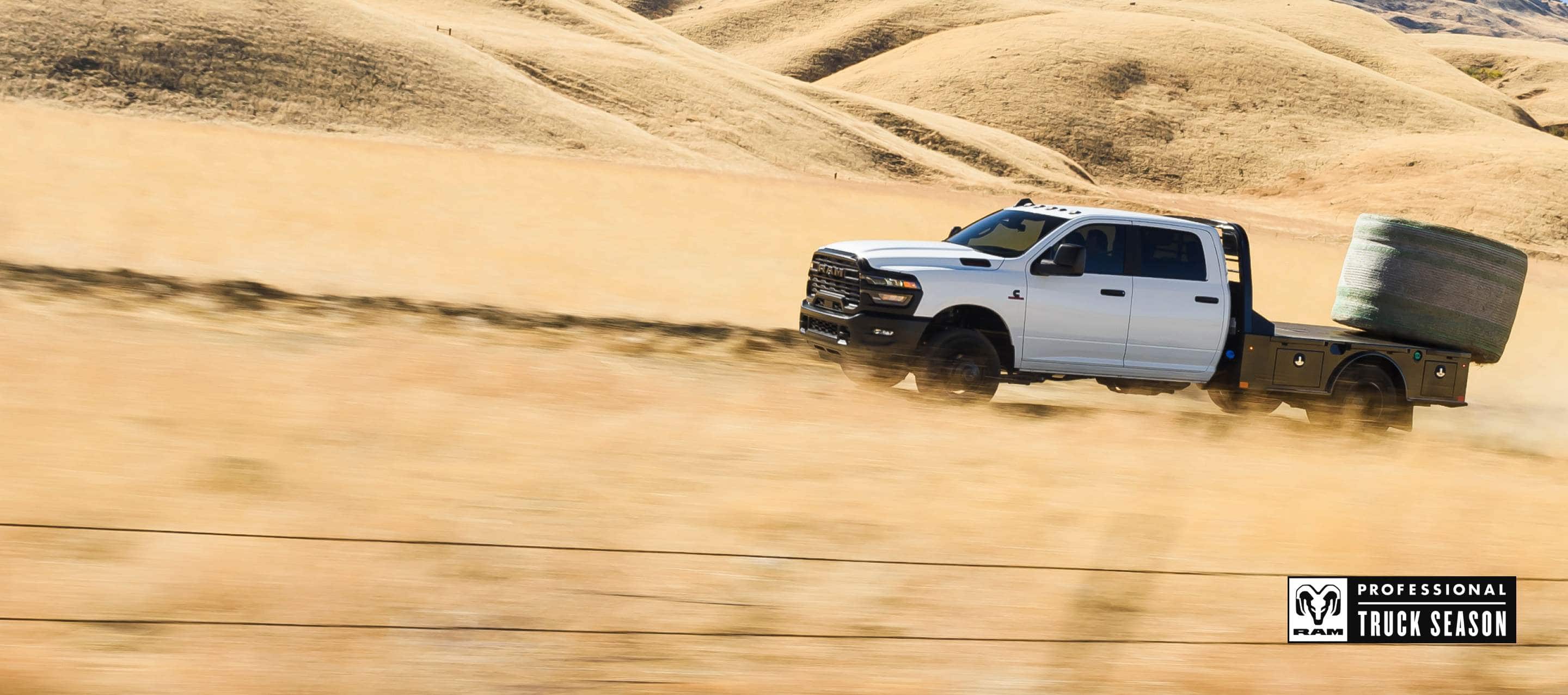 A white 2025 Ram 3500 Tradesman Crew Cab Chassis Cab climbing a sand dune in the desert, with a huge bale of hay on its platform upfit. Ram Professional Truck Season.