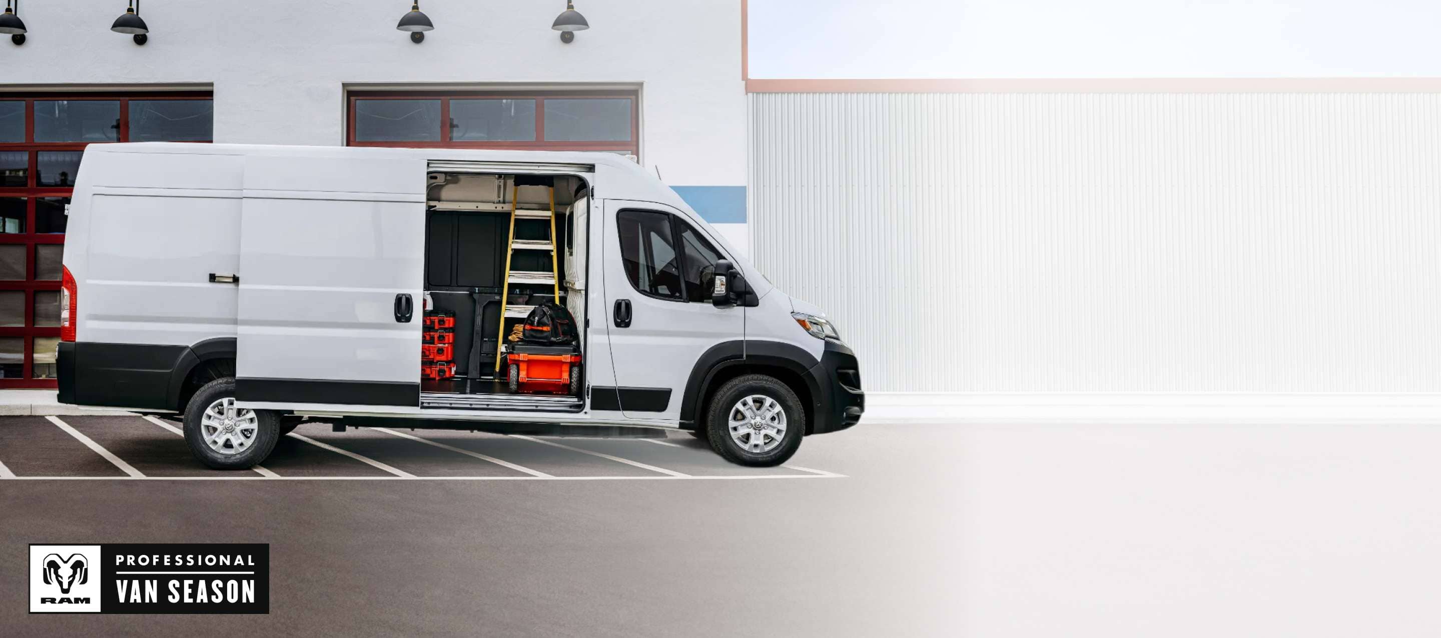 A passenger-side profile of a white 2025 Ram ProMaster EV 3500 SLT High Roof Cargo Van parked beside a commercial garage with its sliding side door open, revealing tools, equipment and a ladder inside. Ram Professional Van Season.
