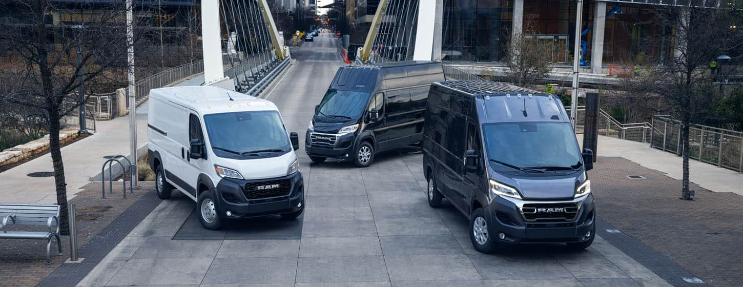 Three 2025 Ram ProMaster models parked just beyond a bridge in the center of a large city, from left to right: a white Ram ProMaster 1500 Tradesman Cargo Van Standard Roof, a black Ram ProMaster 3500 SLT Plus Cargo Van Super High Roof and a black Ram ProMaster 3500 SLT Cargo Van High Roof.