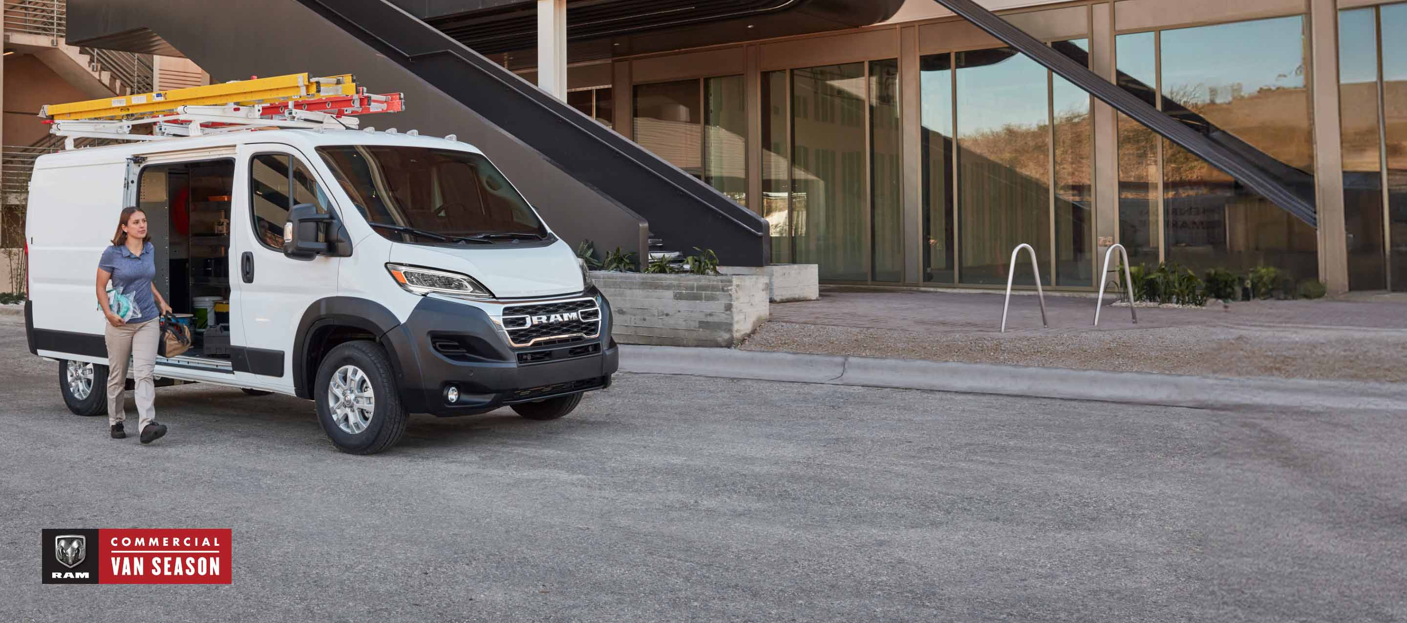 A passenger-side front angle of a white 2025 Ram ProMaster SLT Standard Roof Cargo Van, parked next to a contemporary building with two ladders on its roof rack and its passenger-side sliding door open. Ram Commercial Van Season.