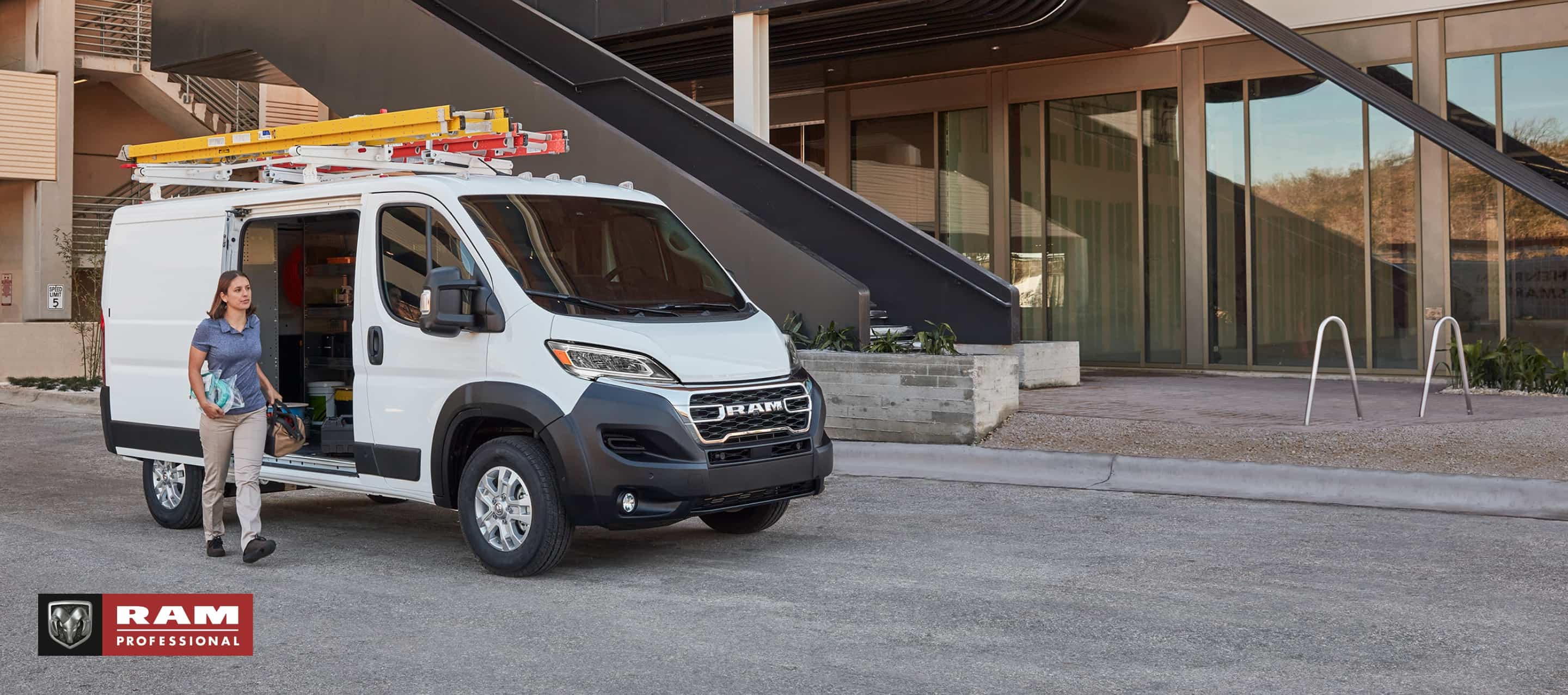 A passenger-side front angle of a white 2025 Ram ProMaster SLT Standard Roof Cargo Van, parked next to a contemporary building with two ladders on its roof rack and its passenger-side sliding door open. Ram Professional.