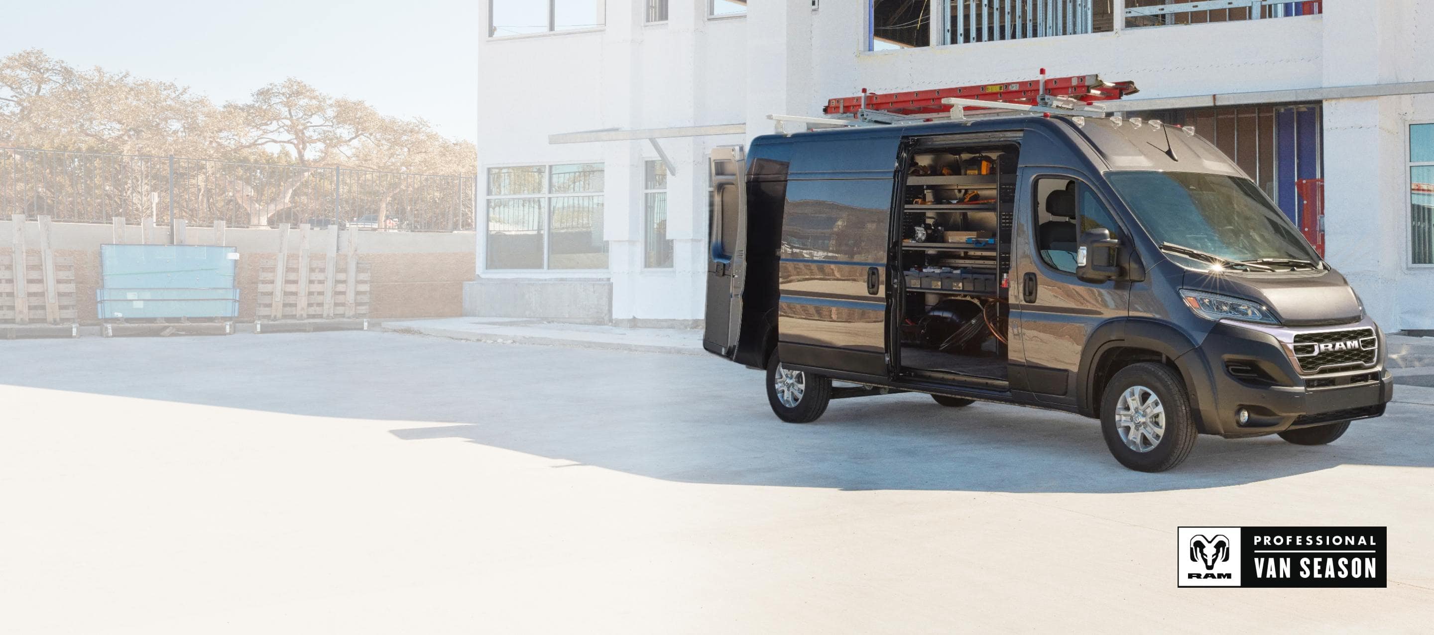 A gray 2025 Ram ProMaster 3500 SLT High Roof Cargo Van parked beside a commercial building with a ladder strapped to its roof rack, its rear doors and sliding side door open revealing shelving full of tools. Ram Professional Van Season.
