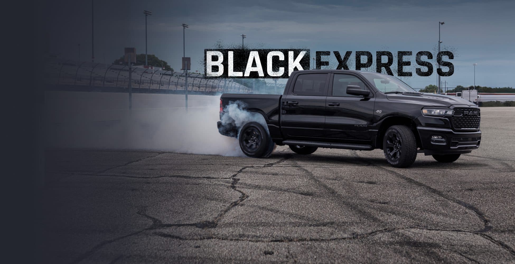 An angled passenger-side profile of a black 2026 Ram 1500 Black Express Crew Cab at a racetrack with smoke billowing from its rear tires. Black Express.