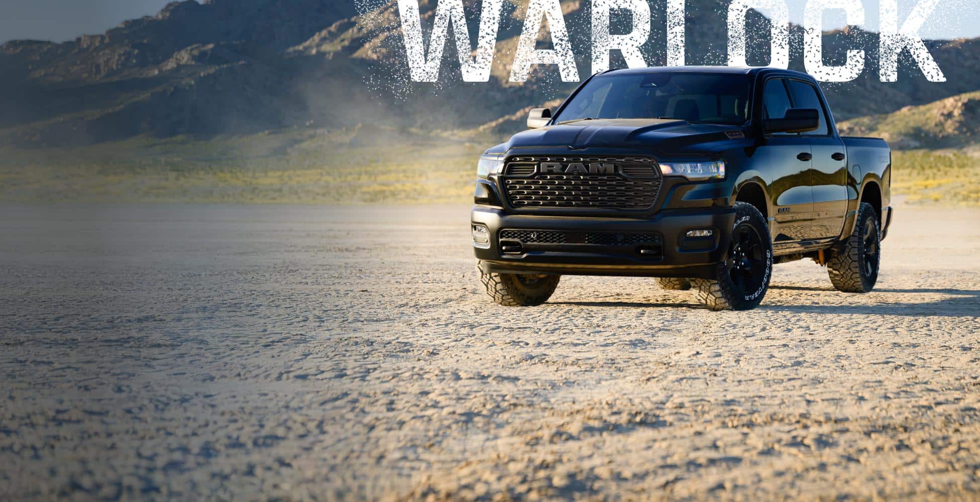 An angled front shot of a black 2026 Ram 1500 Warlock Crew Cab parked on a sandy clearing in the desert. Warlock.