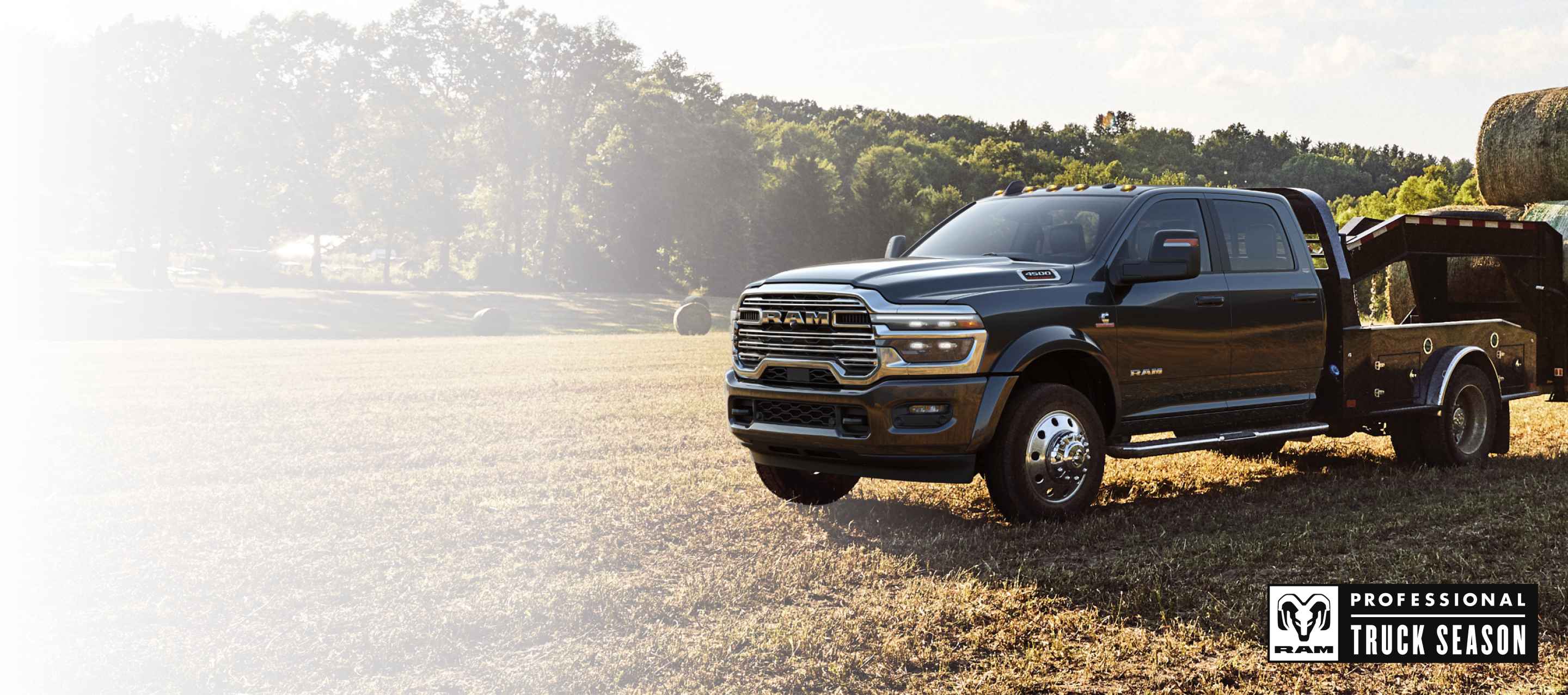A black 2026 Ram 5500 Laramie Chassis Cab Crew Cab with a gooseneck trailer loaded with several bales of hay, parked in an open field. Ram Professional Truck Season.