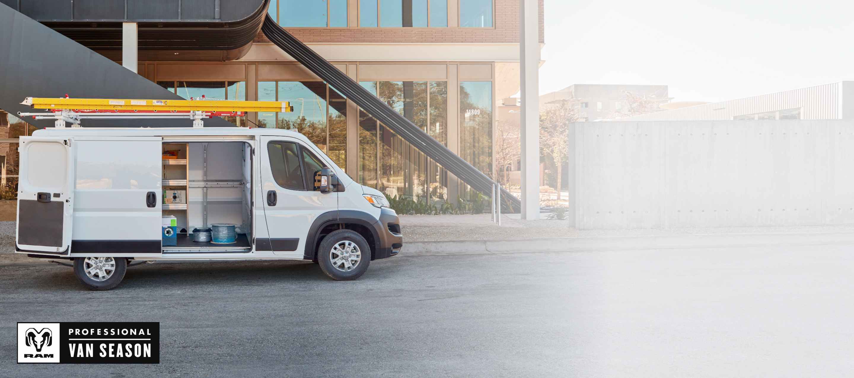 A passenger-side profile of a white 2026 Ram ProMaster 2500 SLT Plus Standard Roof Cargo Van parked next to a contemporary building with a ladder on its roof rack and its passenger-side sliding door and rear doors open. RAM Professional Van Season.
