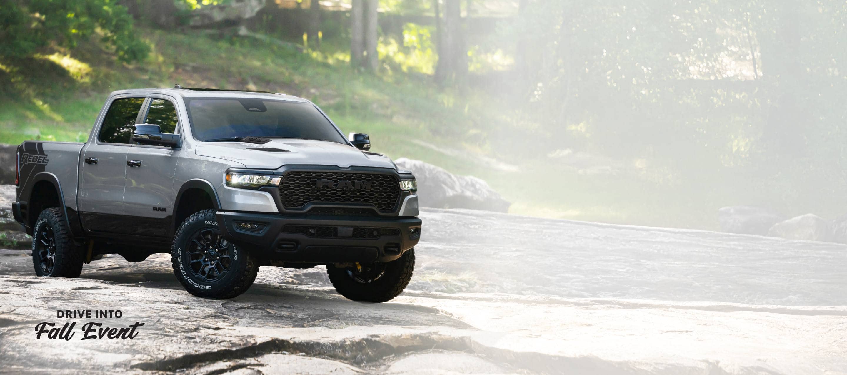 A silver 2026 Ram 1500 Rebel 4x4 Crew Cab traveling across an uneven trail off-road. Drive Into Fall Sales Event.
