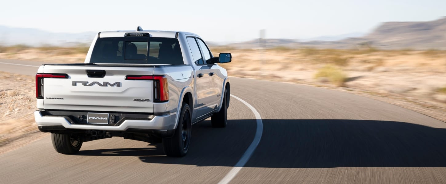 A rear angle of a silver 2027 Ram 1500 Rev Crew Cab traveling down a curved highway in the desert.