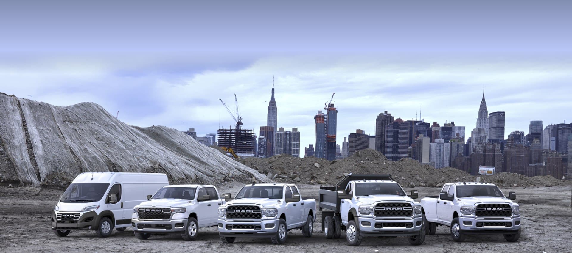 The Ram Brand Lineup in white at a construction site. From left to right: A 2024 Ram ProMaster 3500 SLT High Roof Cargo Van, a 2025 Ram 1500 Tradesman 4x4 Crew Cab, a 2024 Ram 2500 Tradesman 4x4 Crew Cab, a 2024 Ram 5500 Tradesman Chassis Regular Cab with dump body upfit and a 2024 Ram 3500 Tradesman 4x4 Crew Cab.