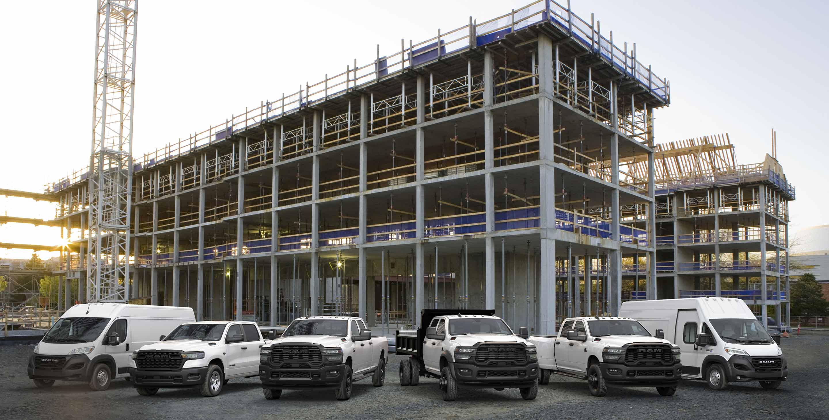 A lineup of 2025 Ram trucks and vans, all in white. From left to right: a Ram ProMaster 3500 Tradesman High Roof Cargo Van, a Ram 1500 Tradesman 4x4 Crew Cab, a Ram 2500 Tradesman 4x4 Crew Cab, a Ram 3500 Tradesman Chassis Cab Crew Cab with a dump body upfit, a Ram 3500 Tradesman 4x4 Crew Cab and a Ram ProMaster EV 3500 Super High Roof Step Van.