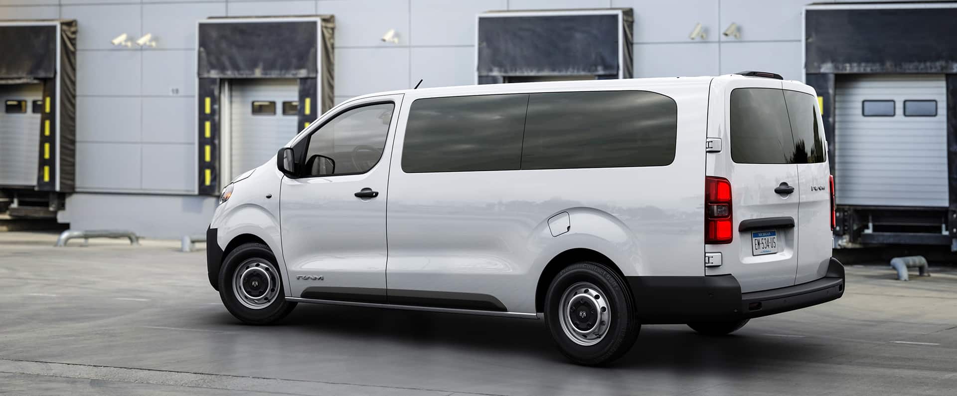 A three-quarter driver side-rear view of a white 2027 Ram ProMaster City® Tradesman Cargo Van with tinted side and rear windows, closed rear barn doors parked next to a loading dock.