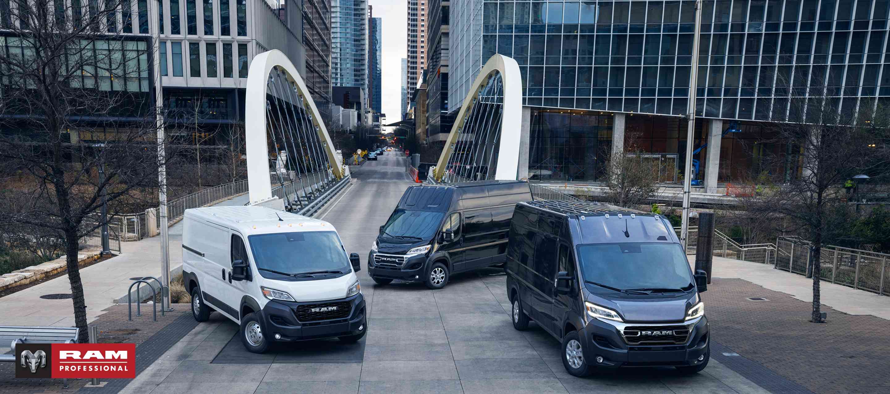 Three 2024 Ram ProMaster models, from left to right: a white Ram ProMaster 1500 Tradesman Standard Roof Cargo Van, a black Ram ProMaster 3500 SLT+ Super High Roof Cargo Van and a black Ram ProMaster 3500 SLT High Roof Cargo Van parked just beyond a bridge in the center of a busy large city. Ram Professional.